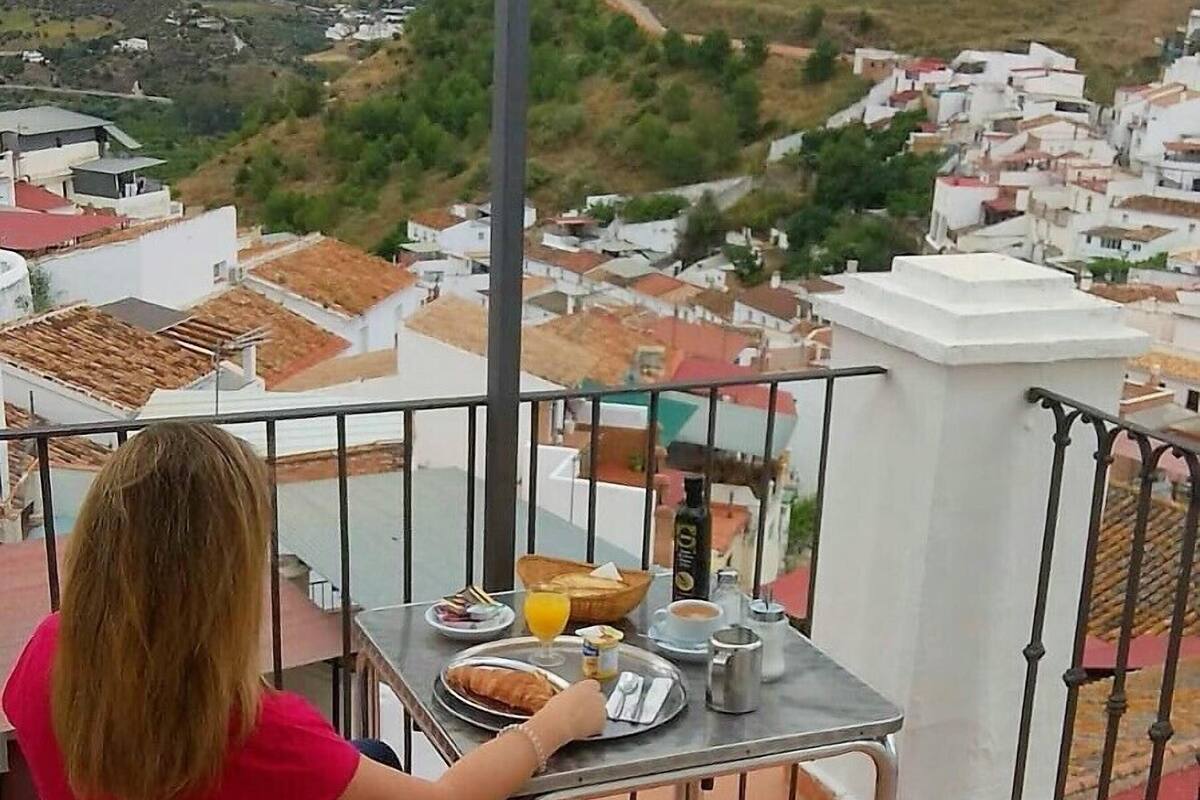 A balcony with a table set for breakfast, overlooking a town with white buildings and a hill in the background.