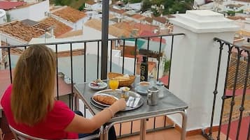 Una persona sentada a una mesa con alimentos para el desayuno en un balcón con vista a un pueblo y colinas.