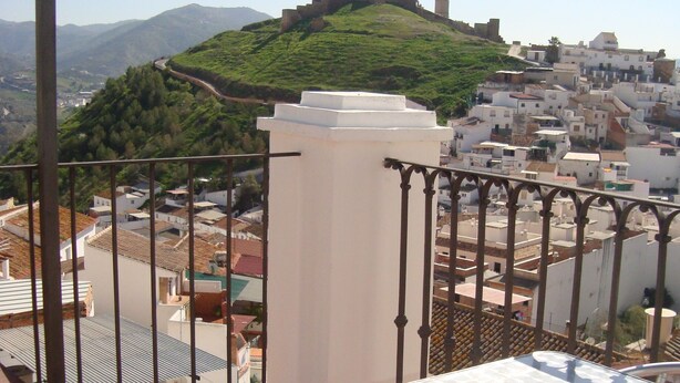 A balcony with a metal chair and table, overlooking a town with white buildings and a hill with a tower. - Hostal Durán (Álora)