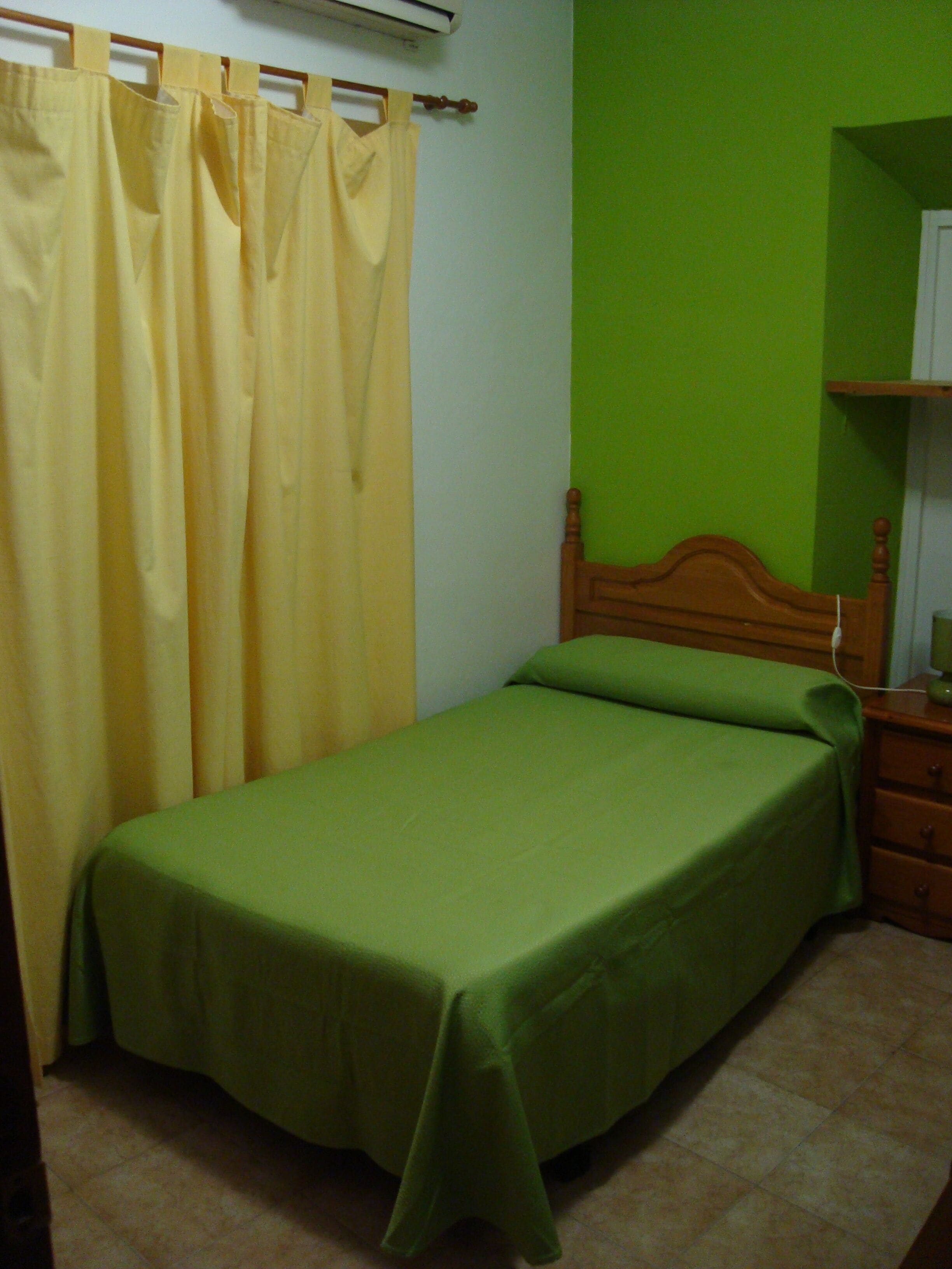 A single bed with a wooden headboard, a green bedspread, and a yellow curtain.