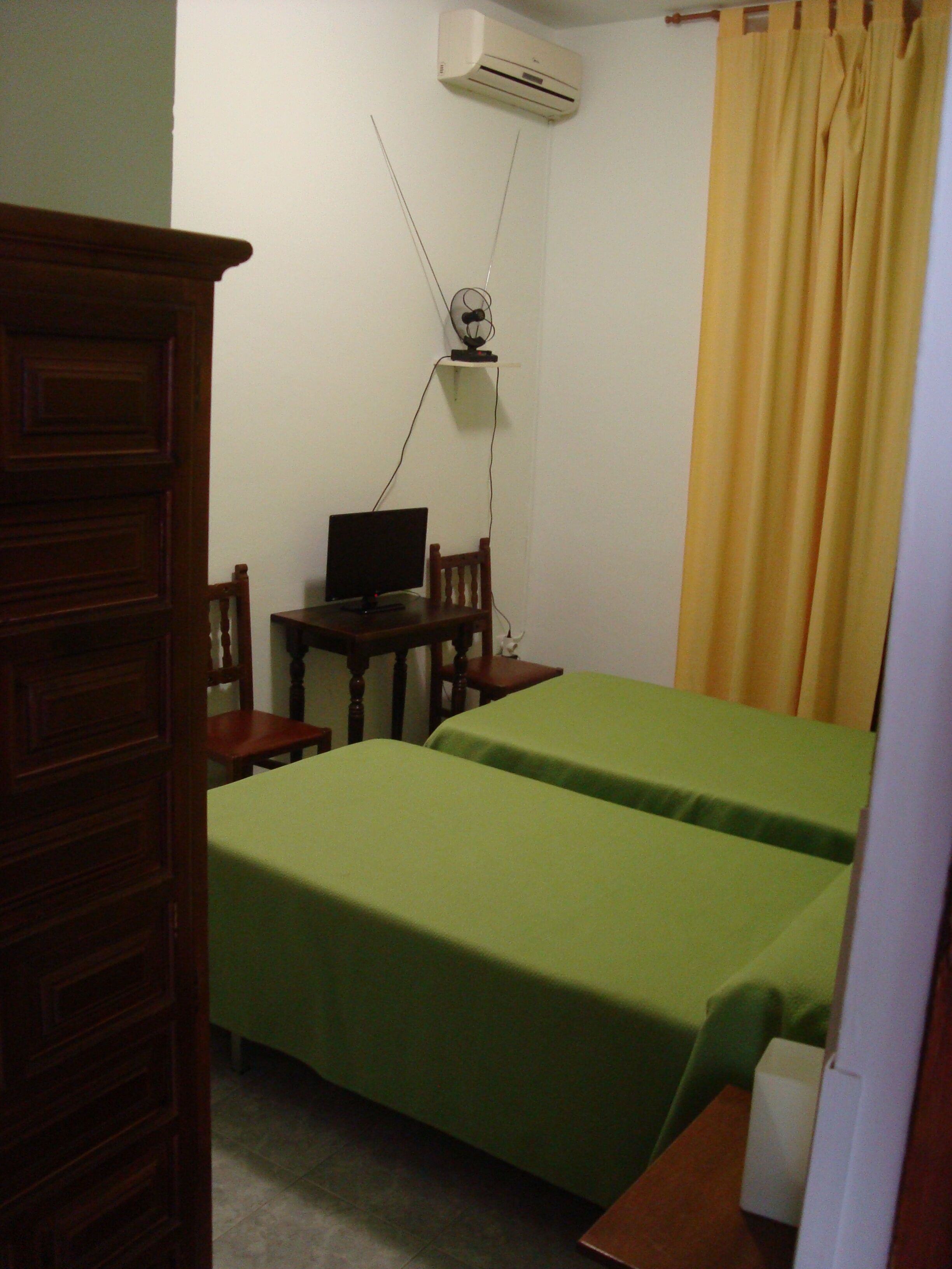 Une chambre avec deux lits, un bureau avec un ordinateur, une commode en bois et un appareil de climatisation fixé au mur.
