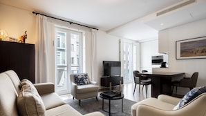 Una sala de estar moderna con un sofá, un sillón y una pequeña mesa de centro. Hay un televisor y un comedor con sillas.