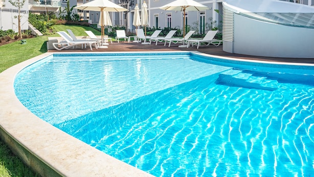 Una piscina con sillas de descanso y sombrillas, rodeada de un césped cuidado y un edificio con balcones.