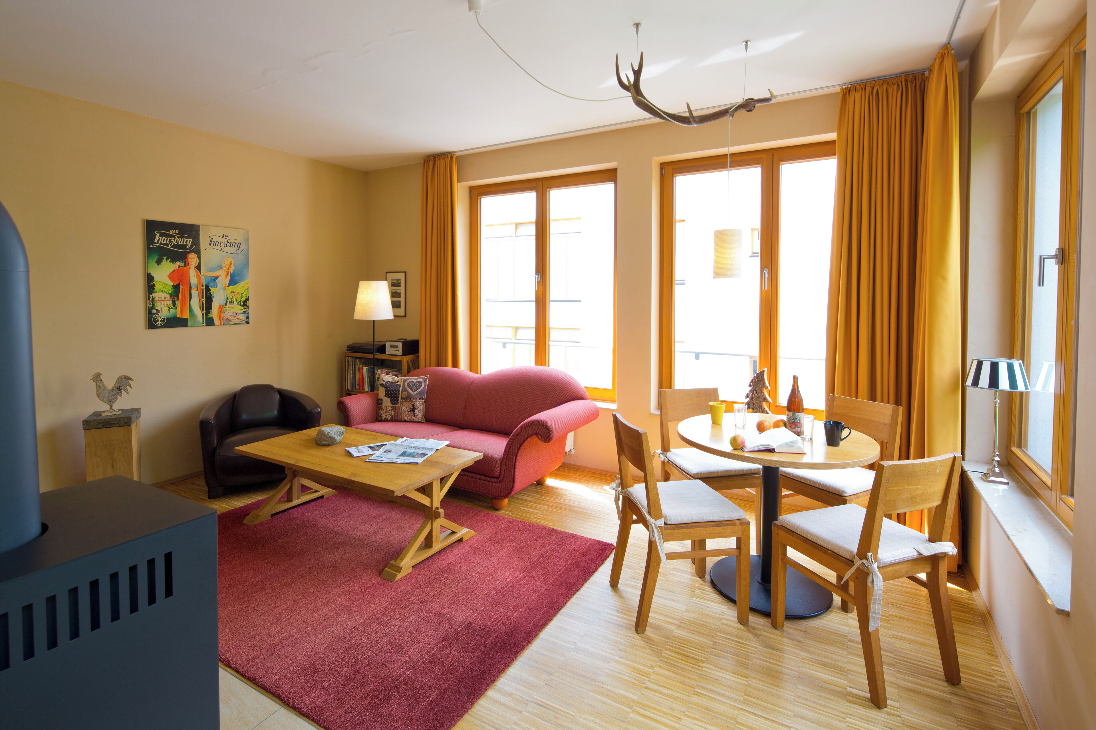 A hotel room with a red sofa, a wooden dining table with chairs, a fireplace, and large windows with curtains.