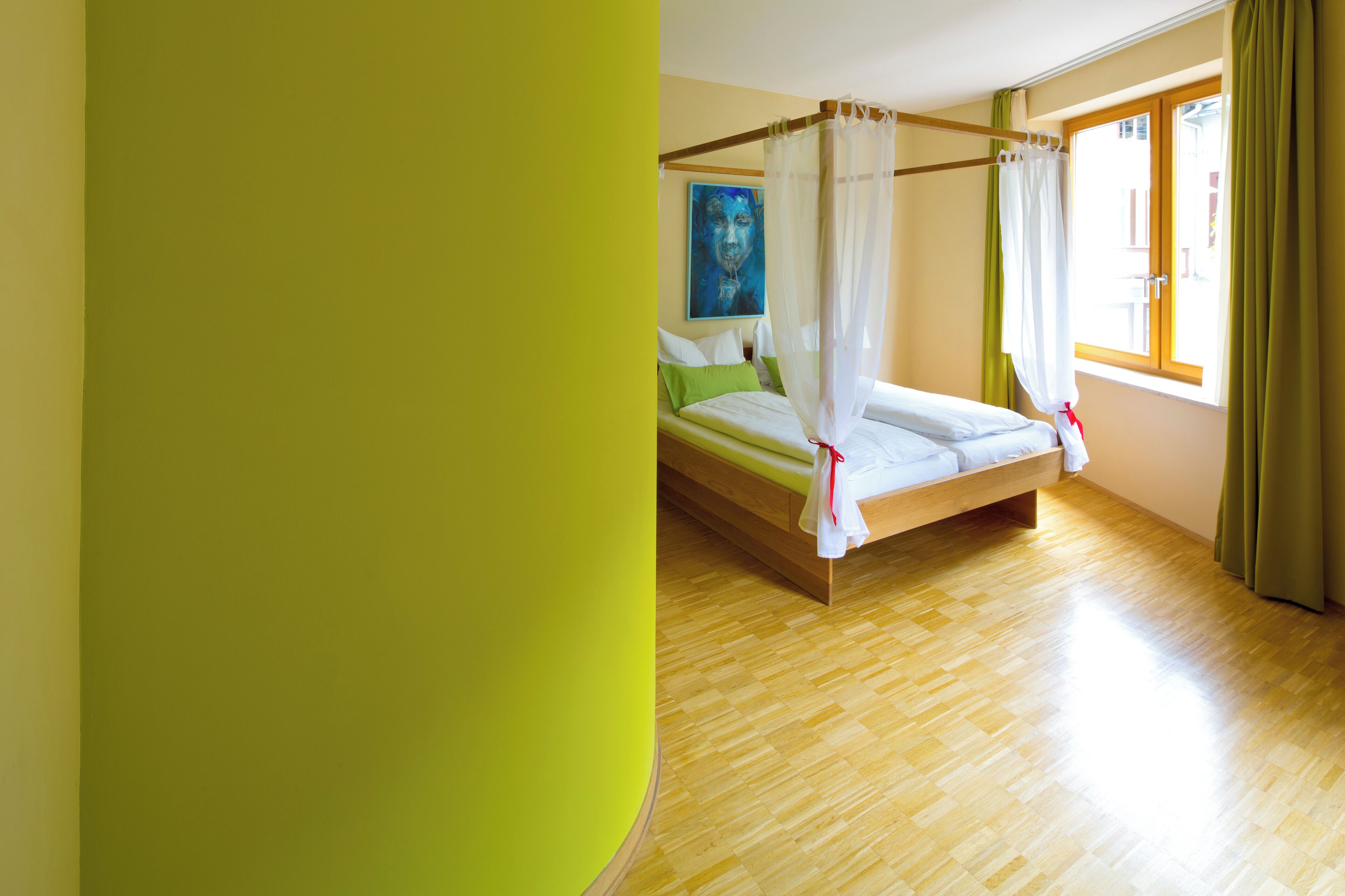 A room with a wooden floor, a bed with a canopy, green pillows, and a painting on the wall.