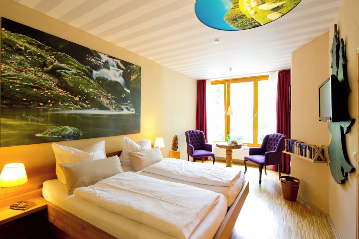 A hotel room with two beds, a large painting of a waterfall, a television, and a bookshelf.