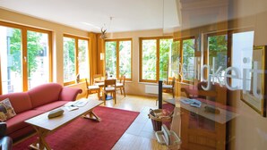 Un salon lumineux avec un canapé rouge, une table basse en bois et une table à manger en verre. De grandes fenêtres offrent une vue sur la verdure environnante.