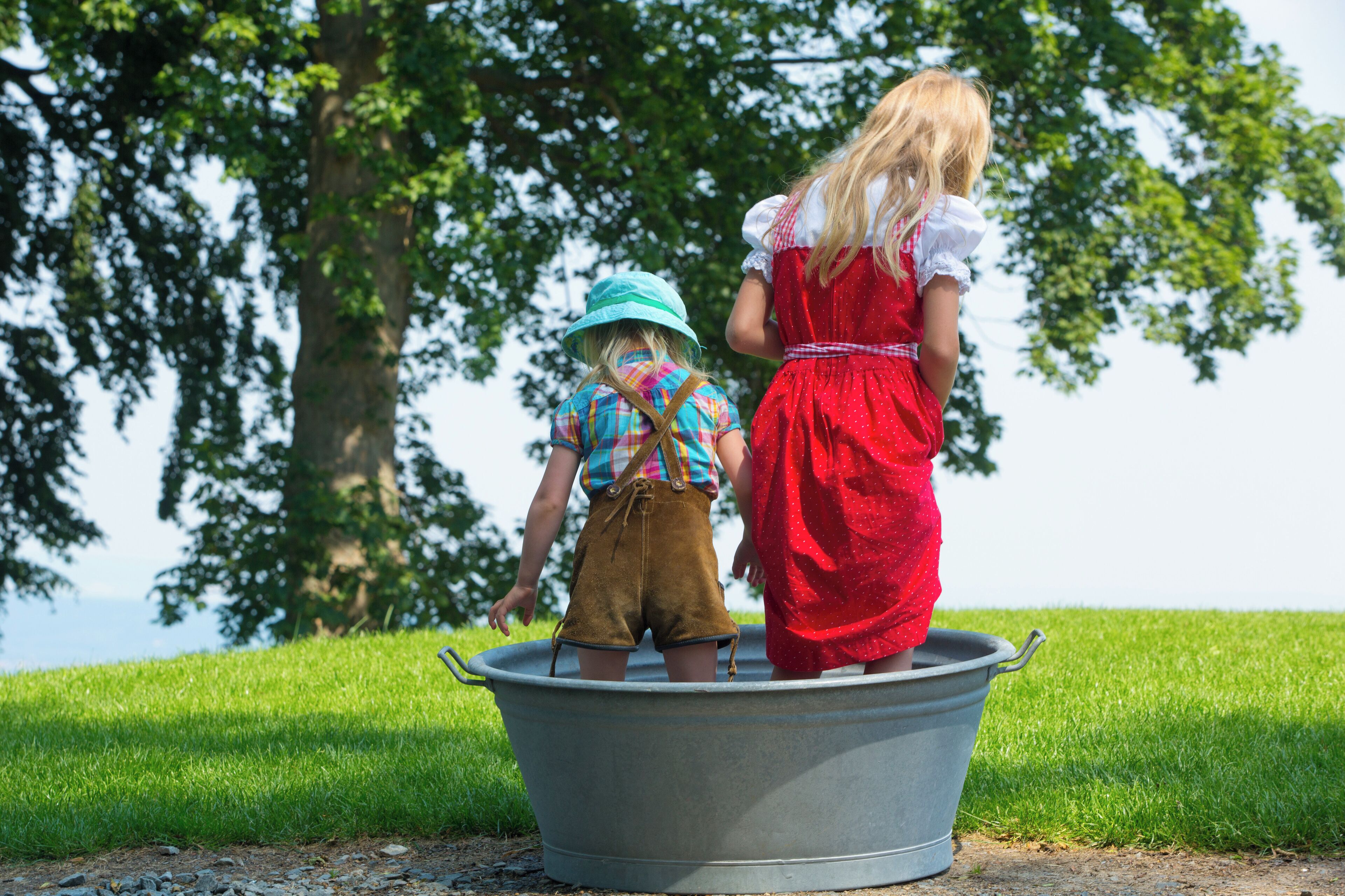 Two children standing in a large metal tub outdoors with grass and trees in the background.