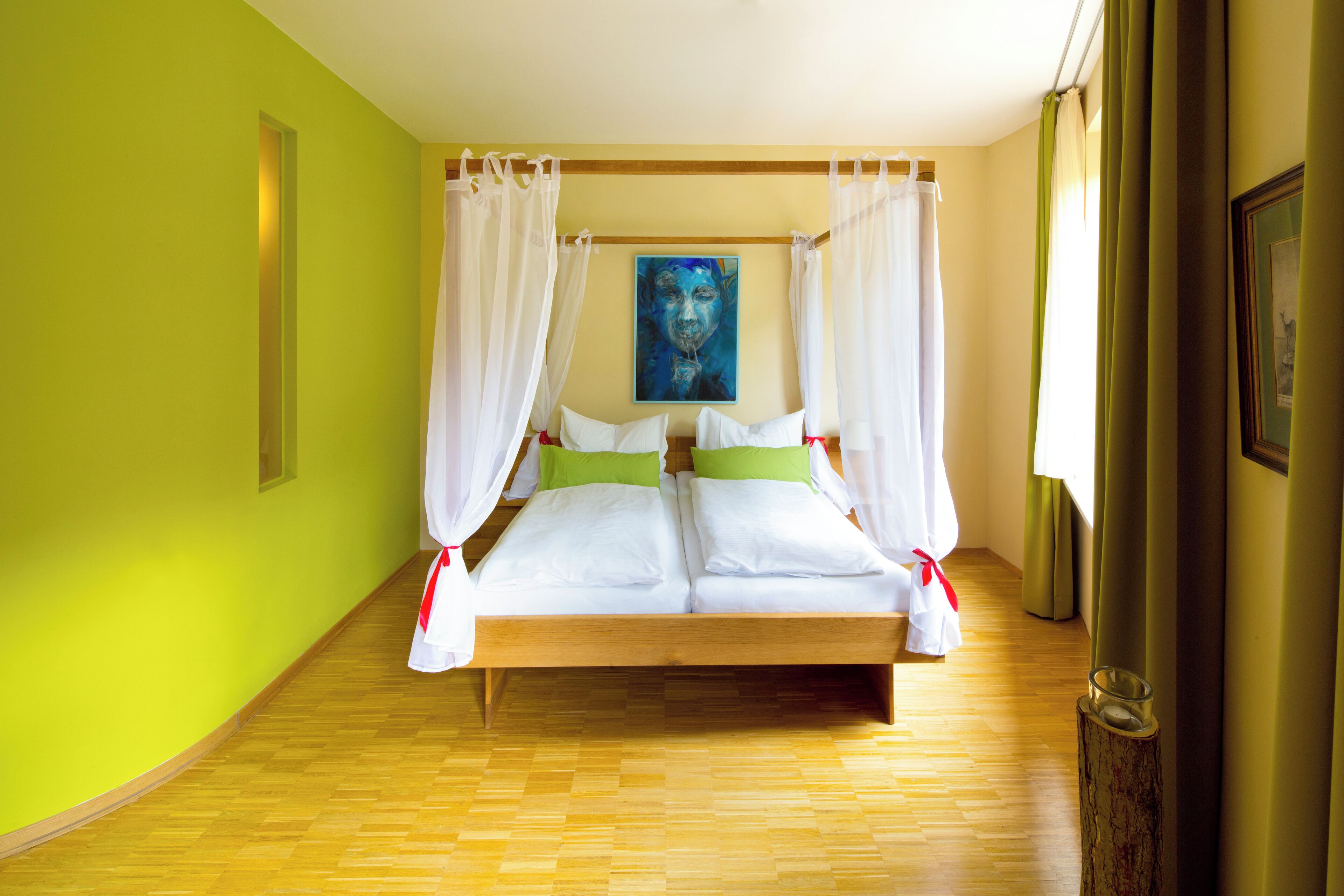 A double bed with white bedding and green pillows, a painting on the wall, and a wooden floor.