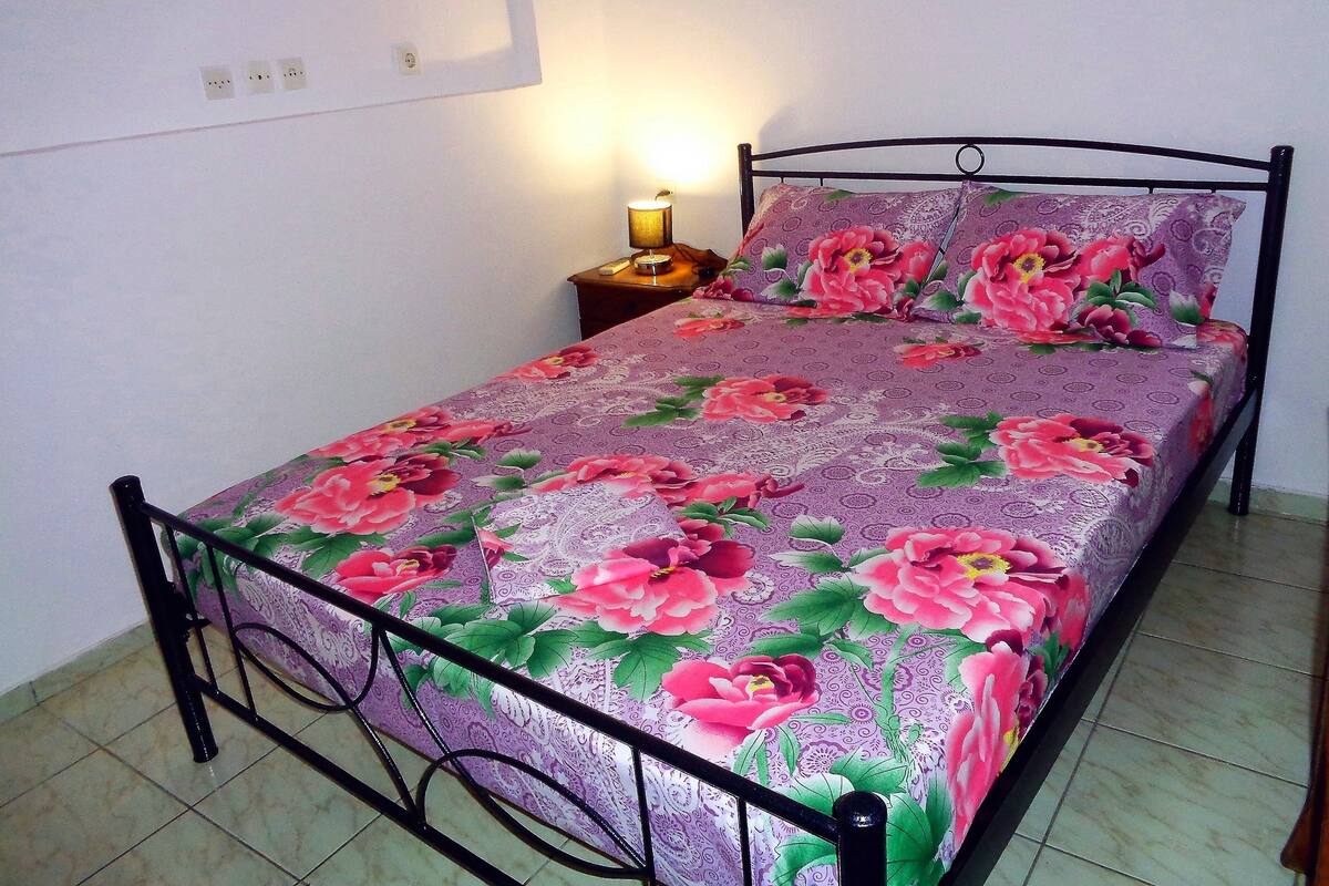 A bed with a floral bedspread and matching pillows, a nightstand with a lamp, and a wall socket.