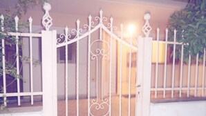 A white metal gate with decorative elements, a white wall, and a plant to the right. - Elena's House (Kalamata)