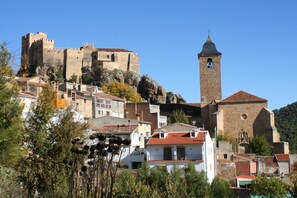 Un pueblo histórico con un castillo destacado en una colina, una iglesia con un campanario y edificios tradicionales con tejados de teja roja.