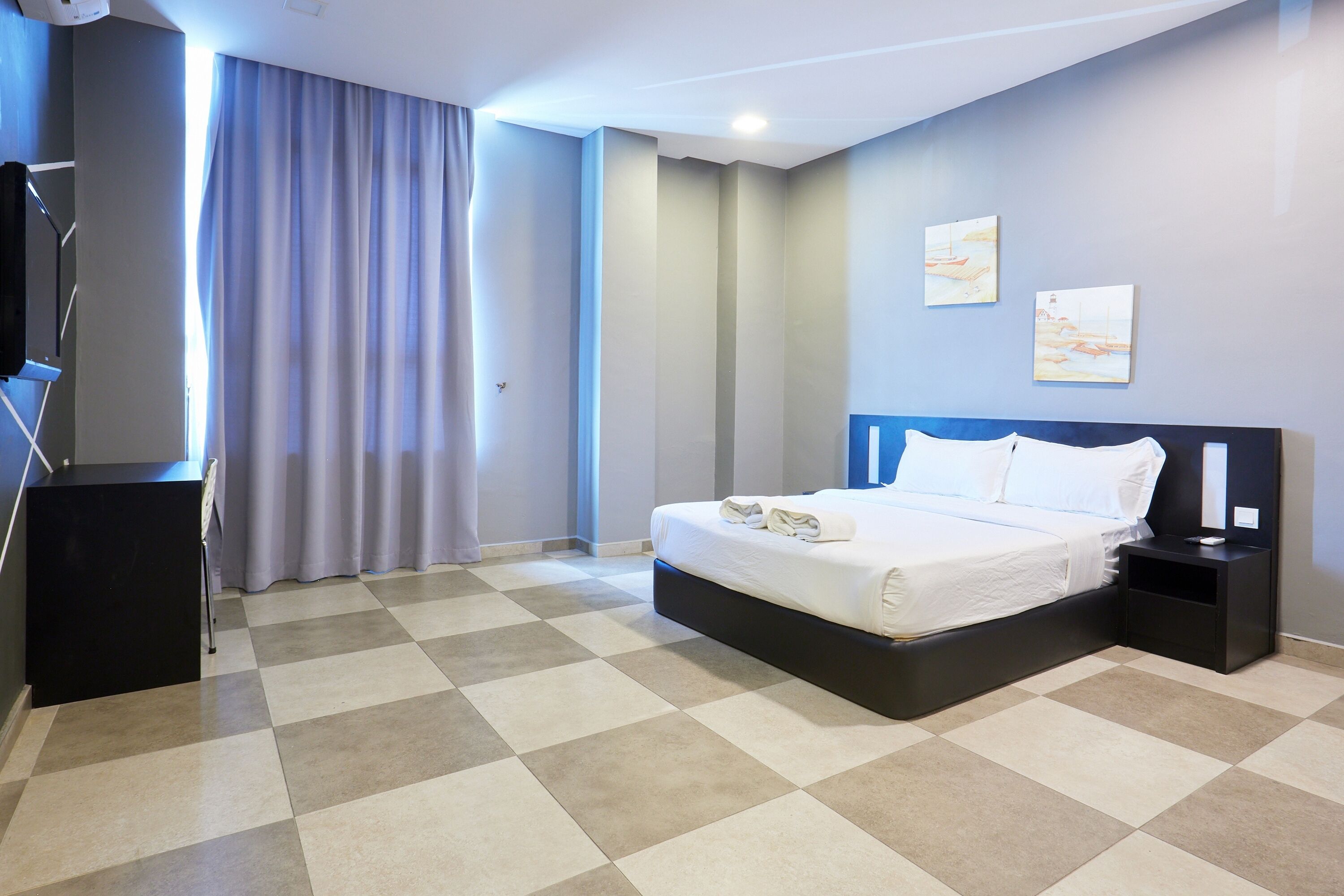 Quarto de hotel com cama grande, mesa de cabeceira, televisão e janela com cortinas.