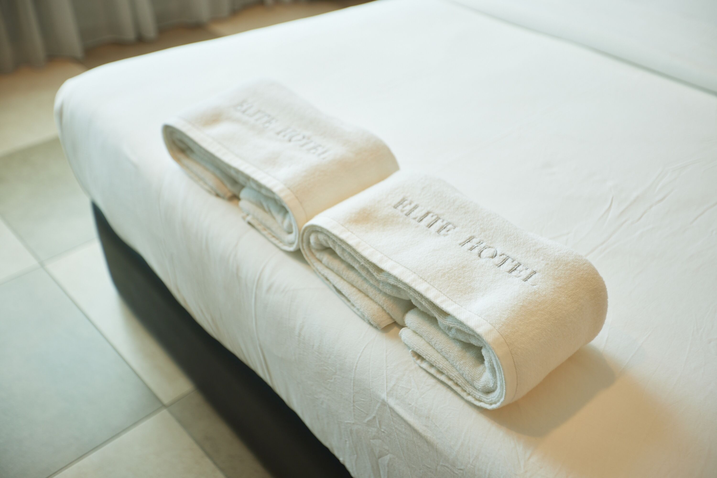 Una cama bien hecha con dos toallas dobladas encima, ambas con la etiqueta 'ELITE HOTEL'.