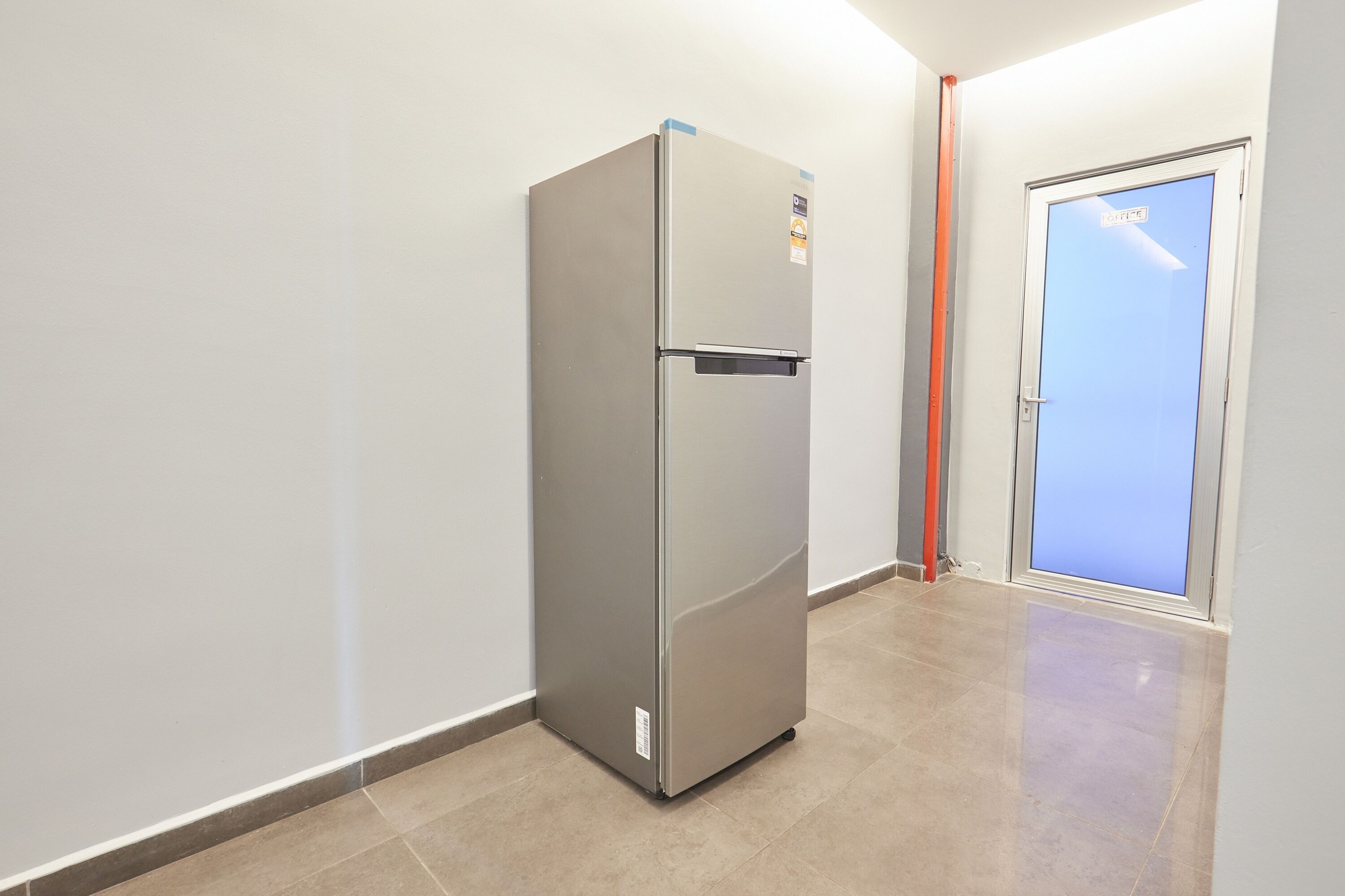 A stainless steel refrigerator in a hallway with a tiled floor and a door with a frosted glass panel.