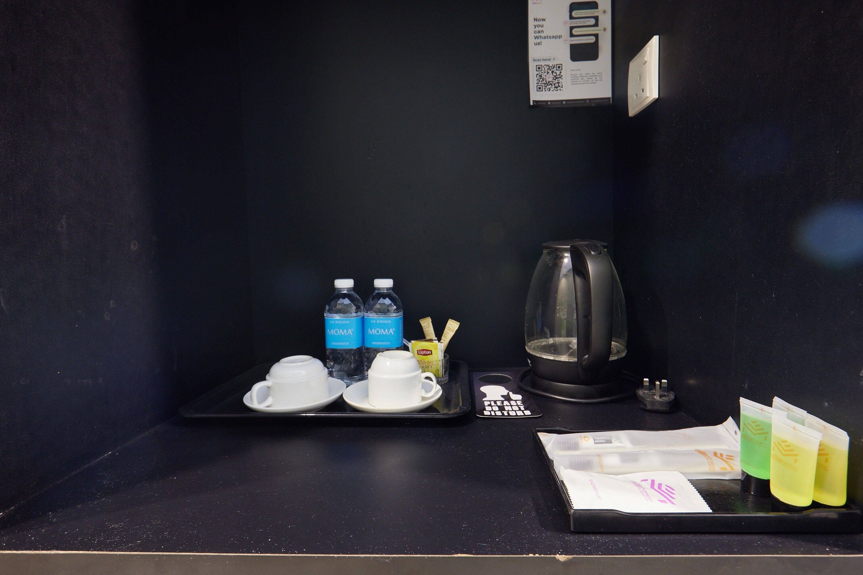 Minibar de una habitación de hotel con una bandeja que contiene dos vasos, dos botellas de agua, una jarra de cristal, una pequeña bandeja con una bolsita de té, una bolsita de té Lipton y varios sobres de té.