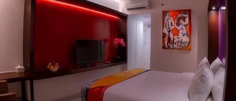 Habitación de hotel con cama, televisión, pared decorada en rojo, un cuadro y escritorio con cesta de fruta.