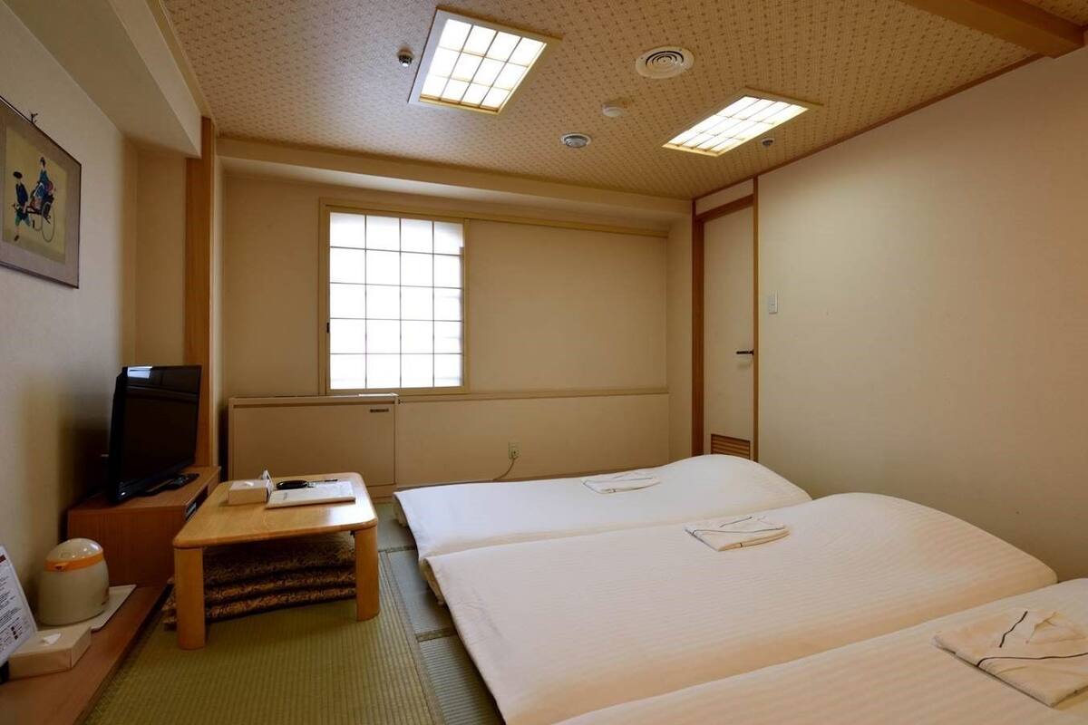 Quarto de hotel com duas camas, uma televisão, uma mesinha e uma janela com persianas.