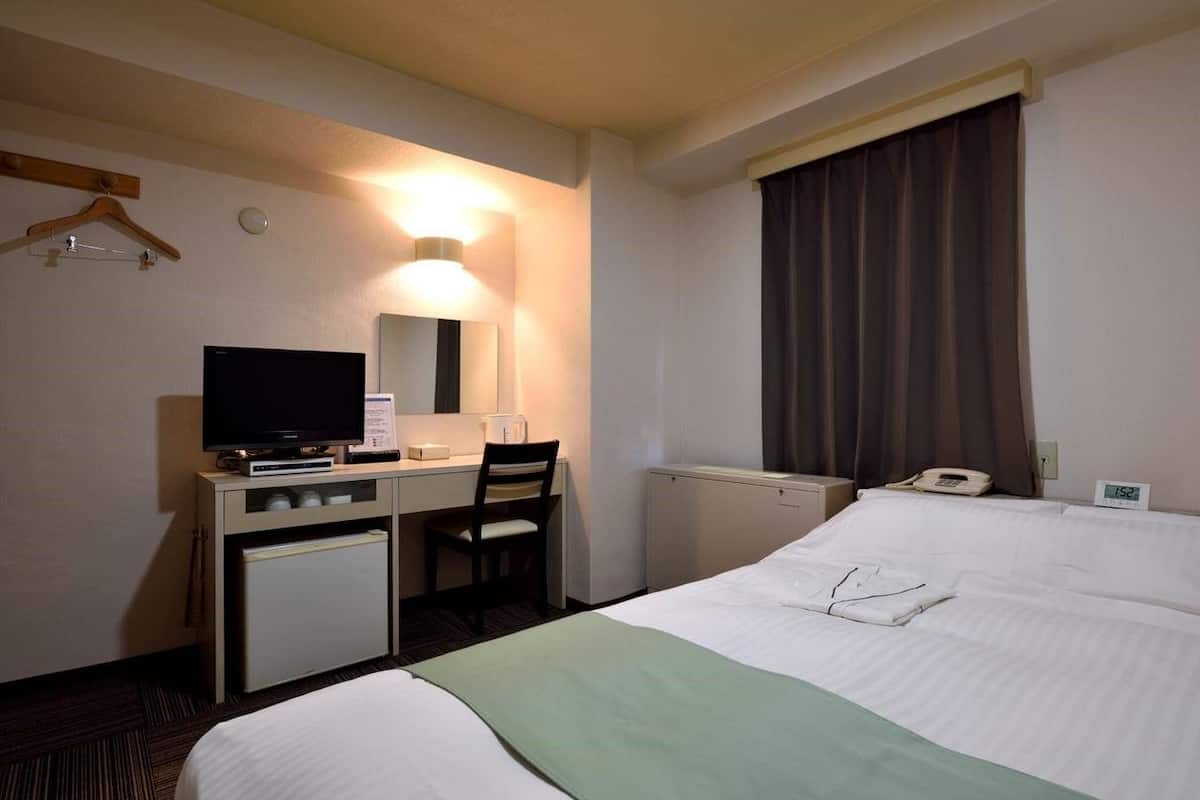 Quarto de hotel com cama, escrivaninha com computador, cadeira, espelho, telefone e relógio.
