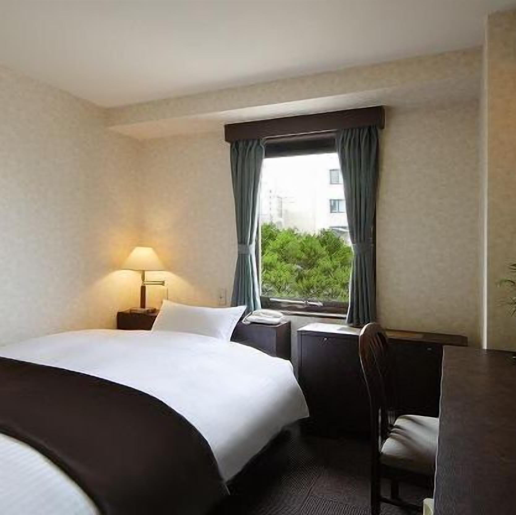 A hotel room with a bed, a desk, a chair, a lamp, and a window with curtains.