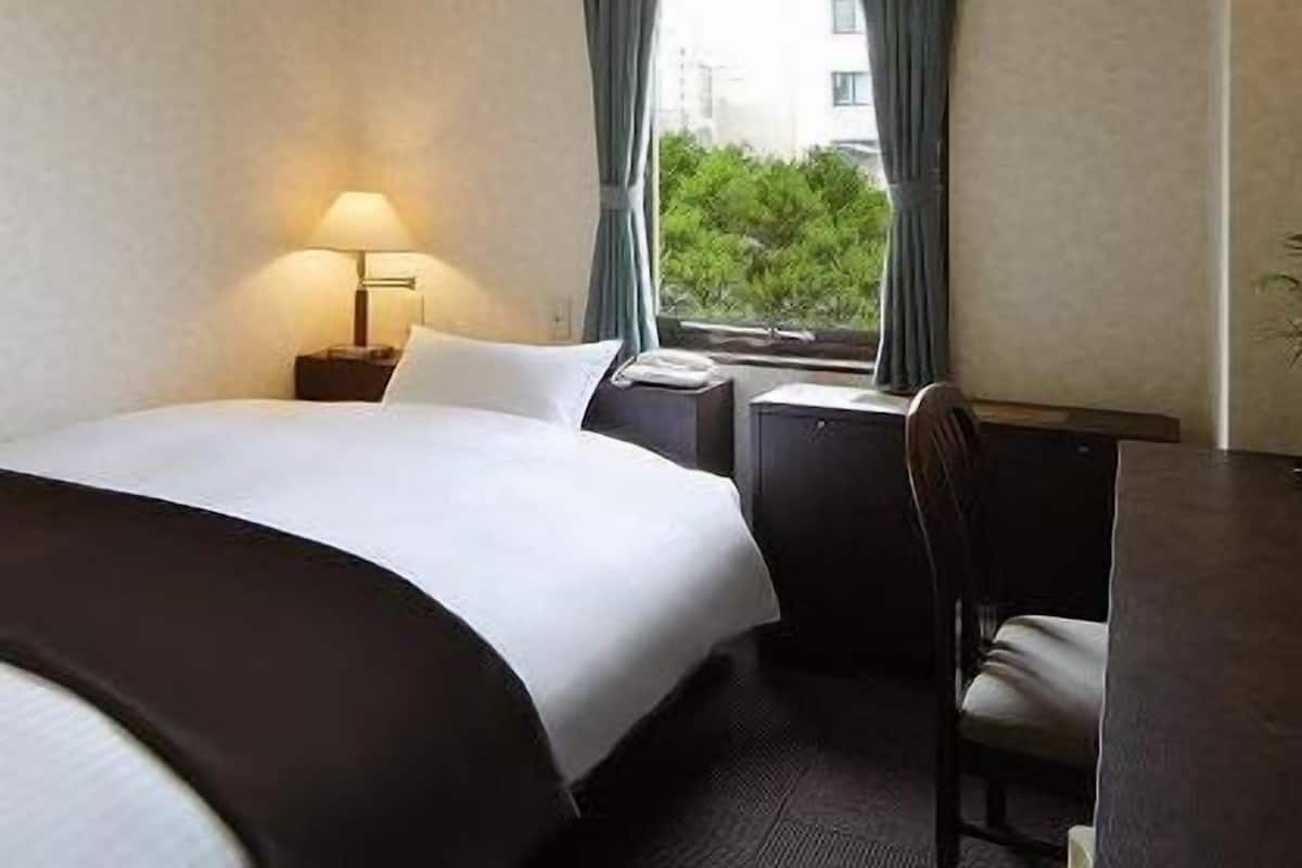 Quarto de hotel com cama, escrivaninha, cadeira, luminária e janela com cortinas.