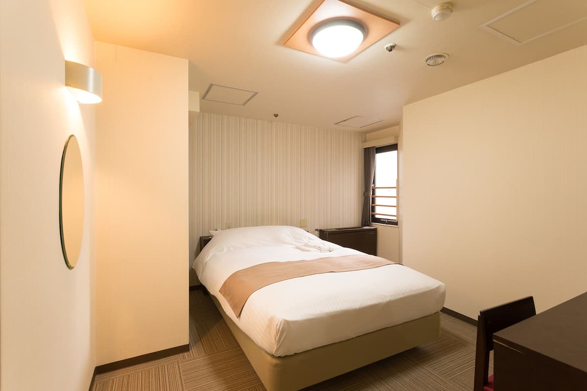 Quarto de hotel com cama, escrivaninha, cadeira, mesa de cabeceira, abajur e janela com persianas.