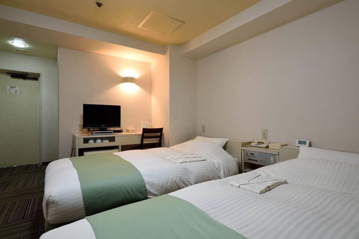 Quarto de hotel com duas camas, uma secretária com televisão, mesa de cabeceira com telefone e uma luminária na parede.