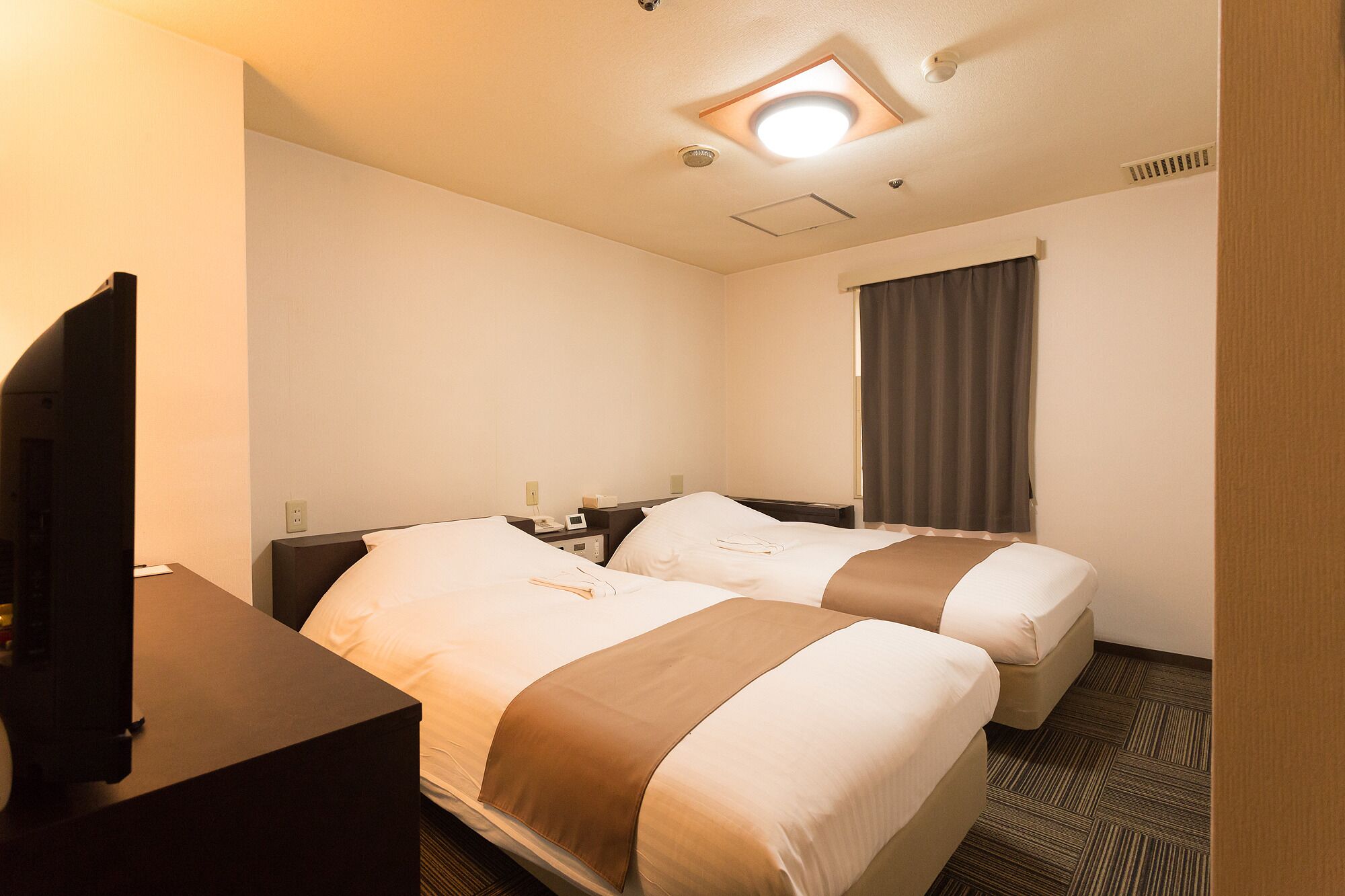 A hotel room with two beds, a desk, a TV, and a window with curtains.