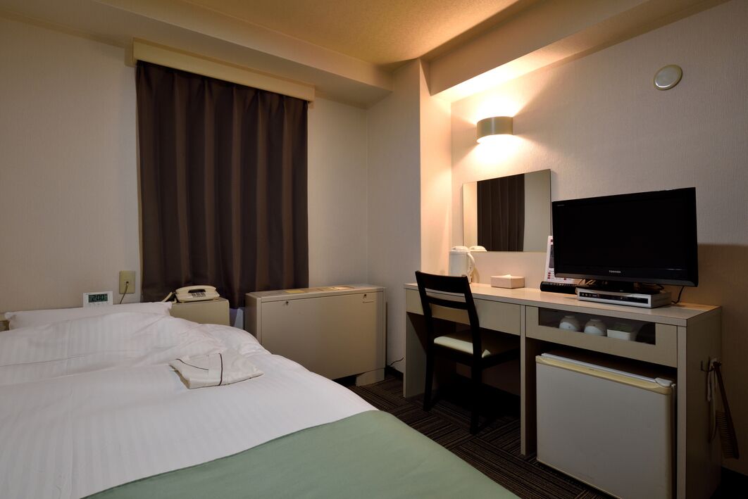 A hotel room with a bed, a desk with a chair, a television, a mirror, and a small refrigerator.