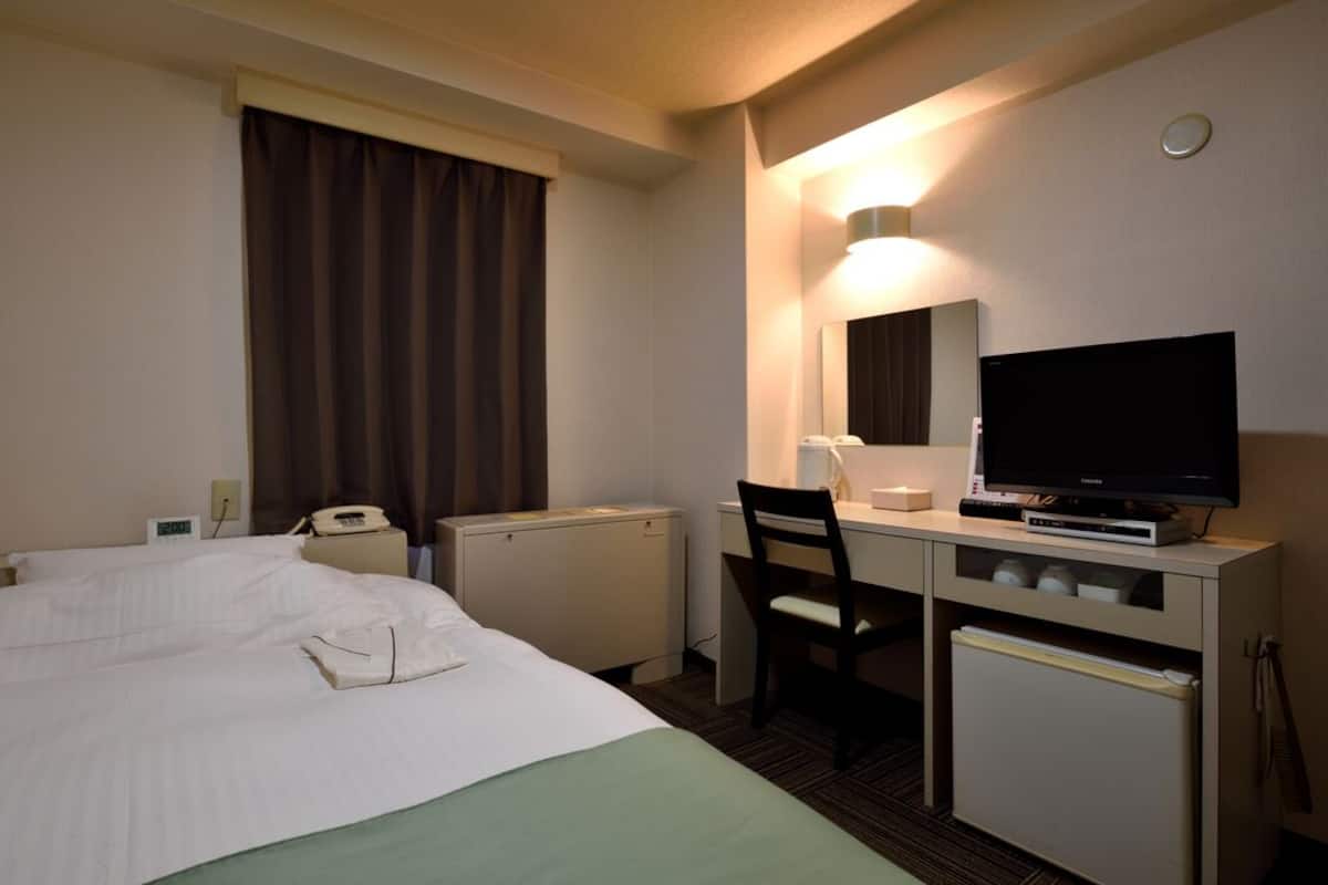 Quarto de hotel com cama, escrivaninha com cadeira, televisão, espelho e pequeno refrigerador.