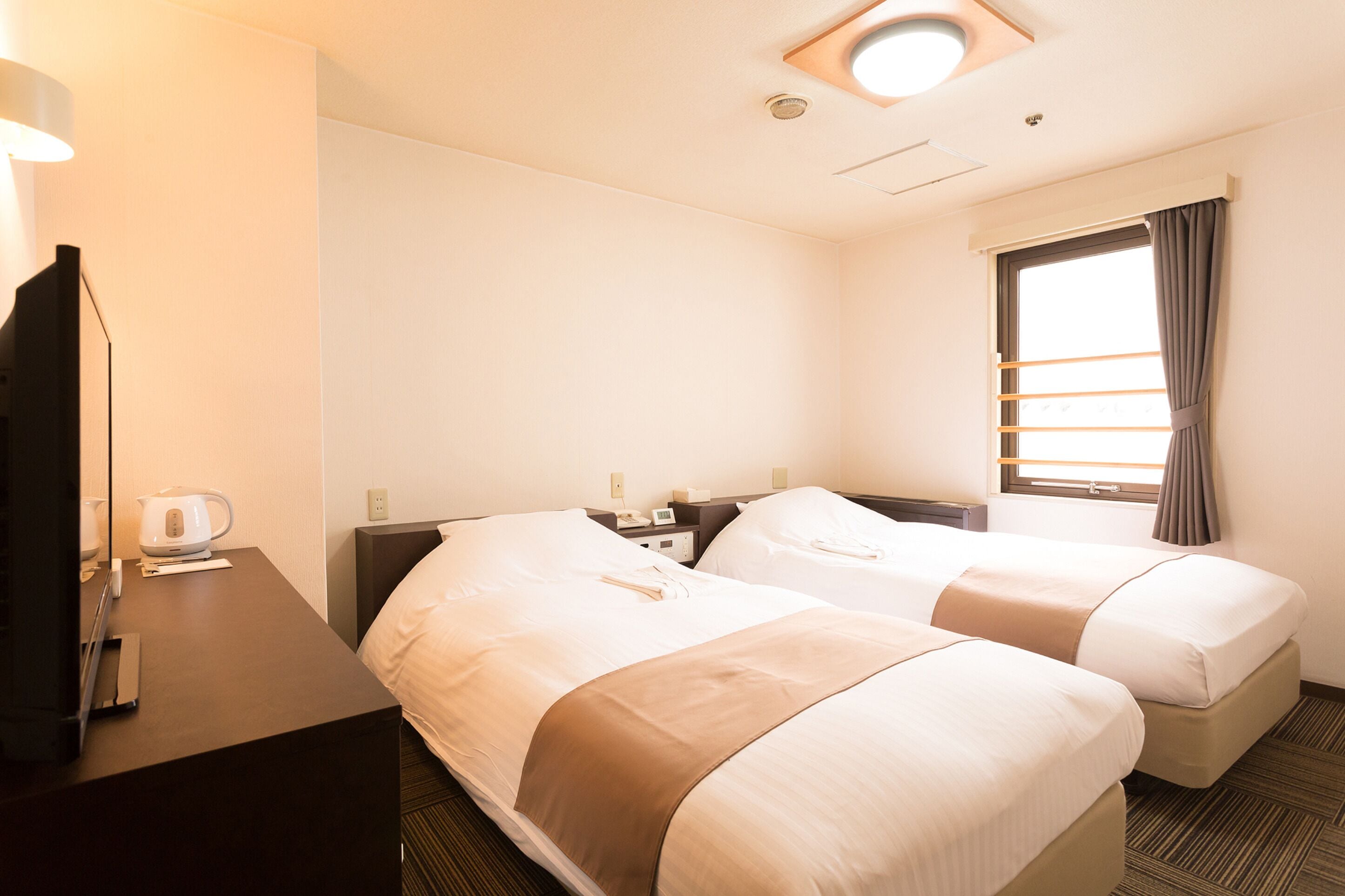 Habitación de hotel con dos camas, un escritorio con lámpara, cafetera y ventana con cortinas.