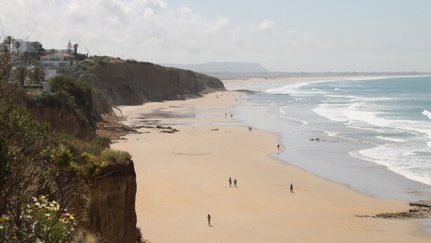 Ein Strand mit sandigem Ufer, eine bewachsene Klippe und einige Spaziergänger.