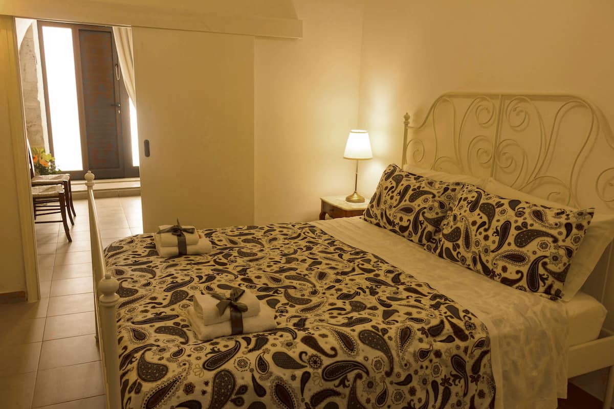A hotel room with a bed, a nightstand with a lamp, a chair, and a view of the outside through a door.