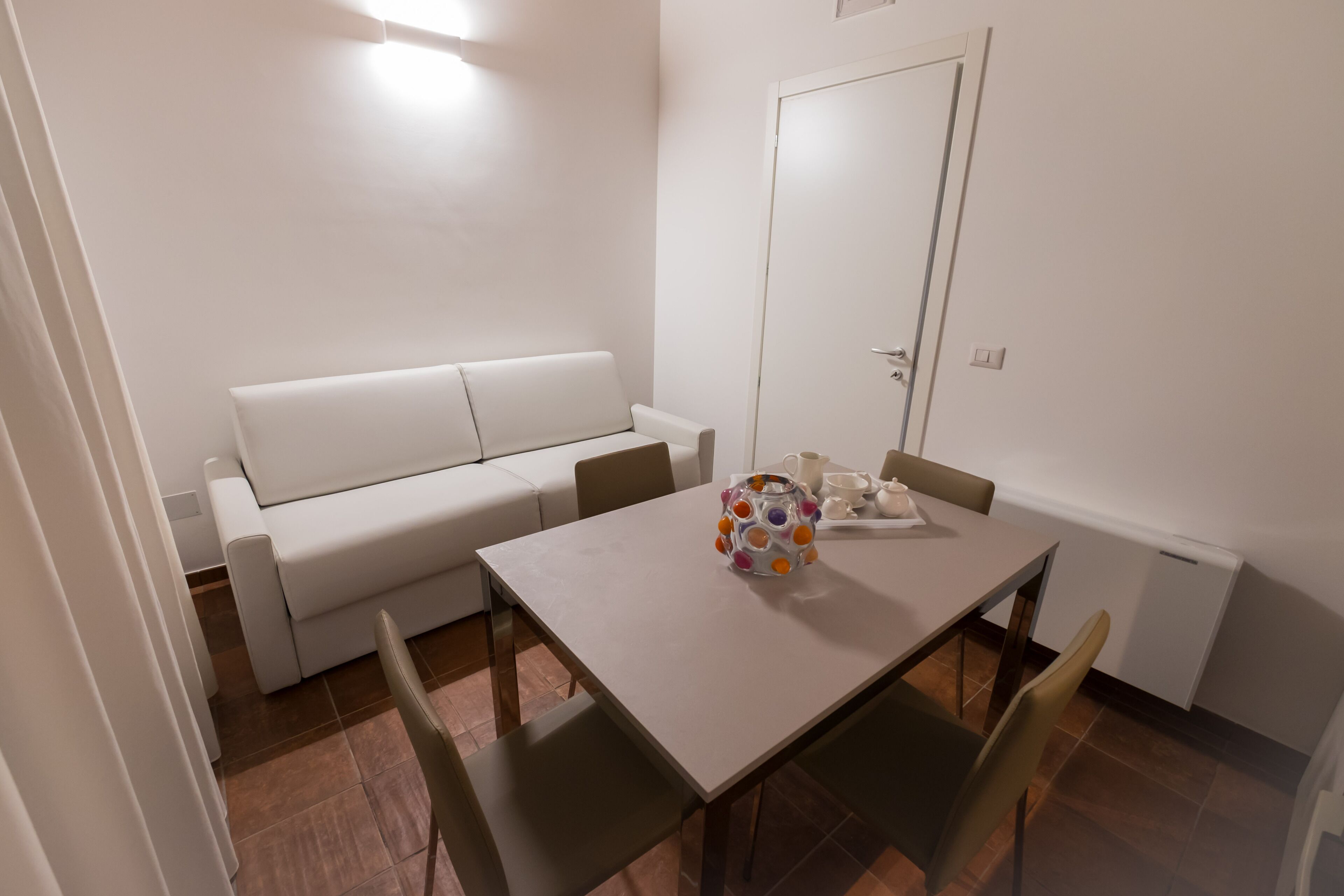 Une petite pièce propre, avec un canapé blanc, une table à manger et quatre chaises, ainsi qu’une porte.
