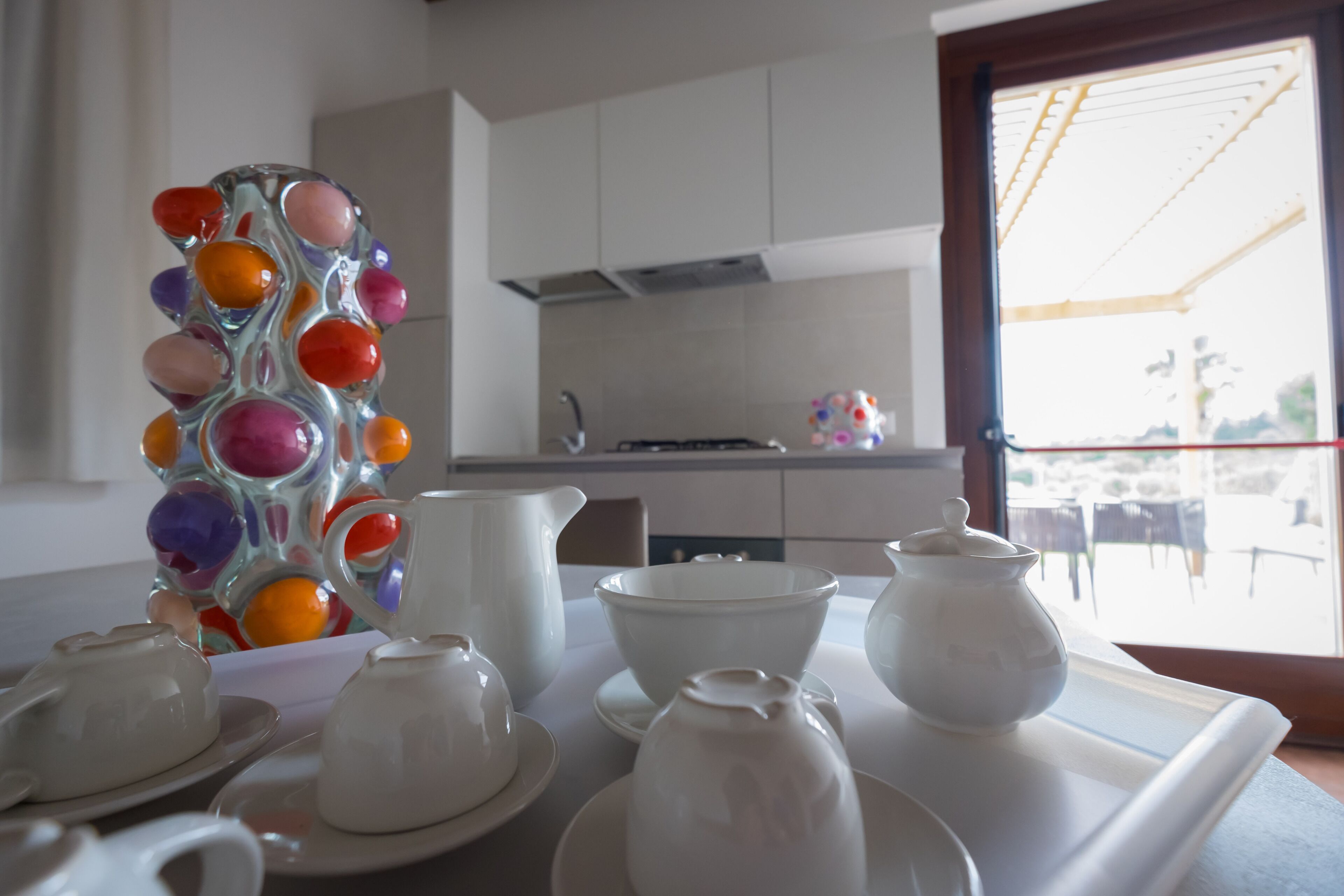 A kitchen with white cabinets, a window with a view of the outside, and a tray with white teacups and a pitcher.