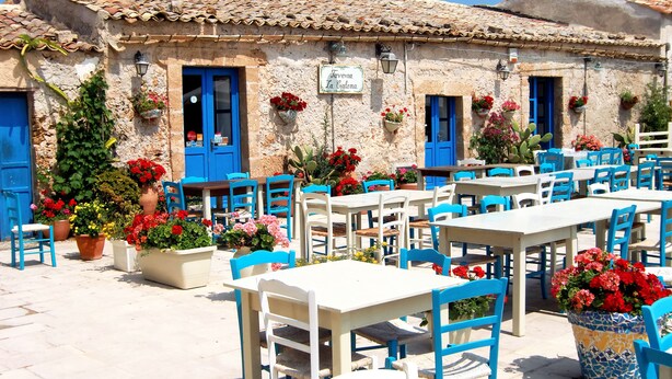 An outdoor café with white tables and blue chairs, surrounded by potted plants and a building with blue doors. - Domus Hyblaea (Palazzolo Acreide)