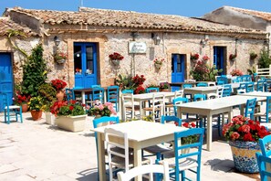 Un café al aire libre con mesas blancas y sillas azules, rodeado de plantas en macetas y un edificio con puertas azules.