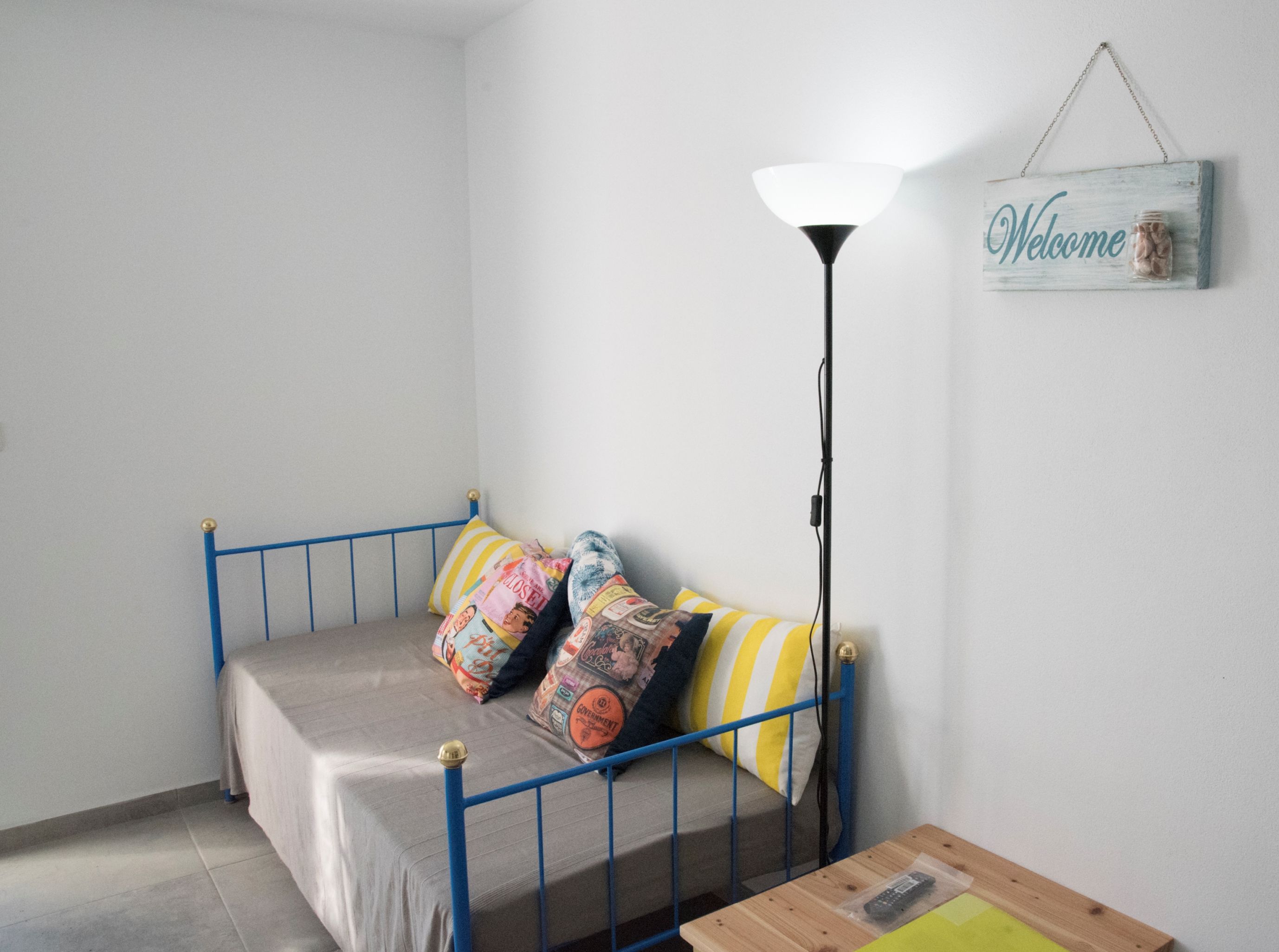 Una piccola camera con un letto singolo, una struttura in metallo blu, un comodino con un telecomando e un cartello con scritto 'Benvenuti' sulla parete.