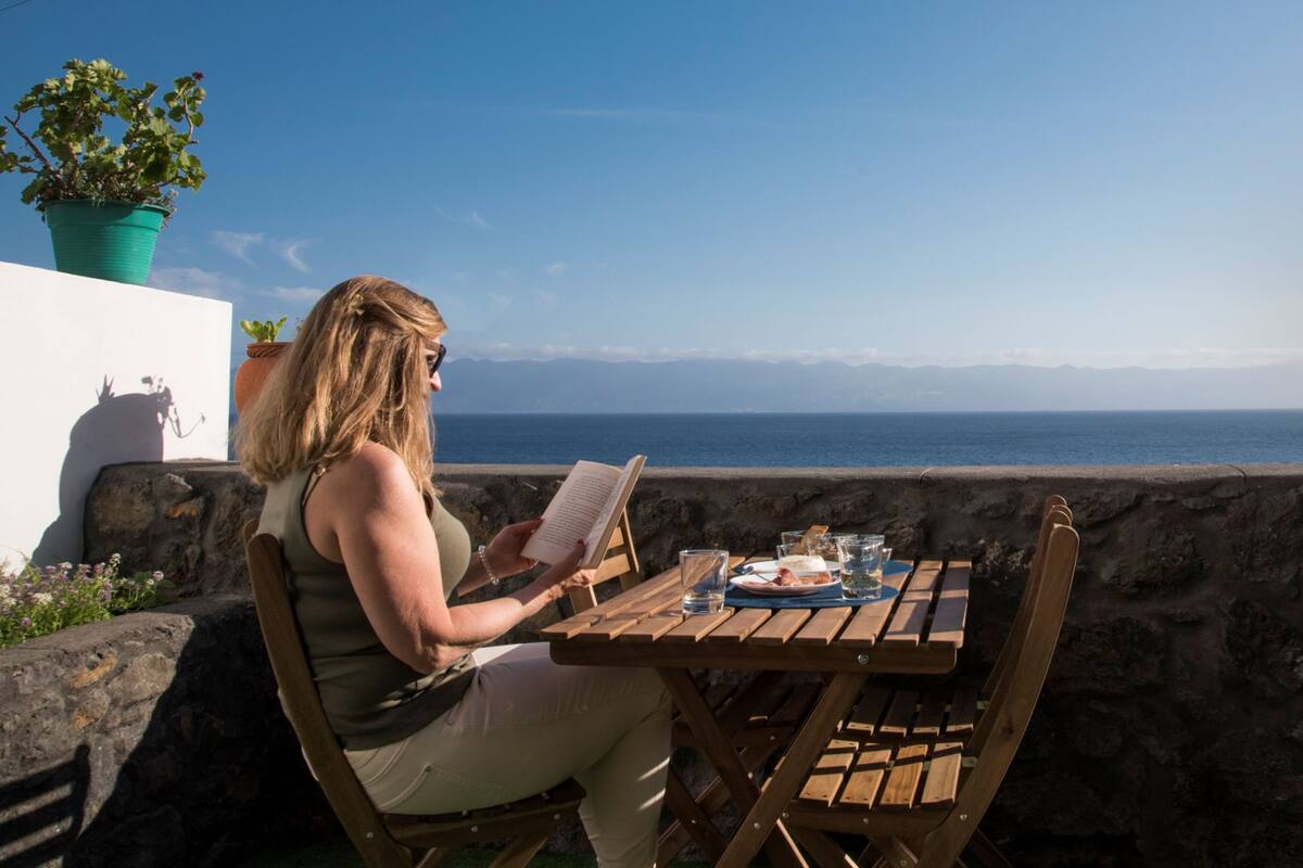 Eine Person liest an einem Holztisch mit Blick auf das Meer und die Berge.