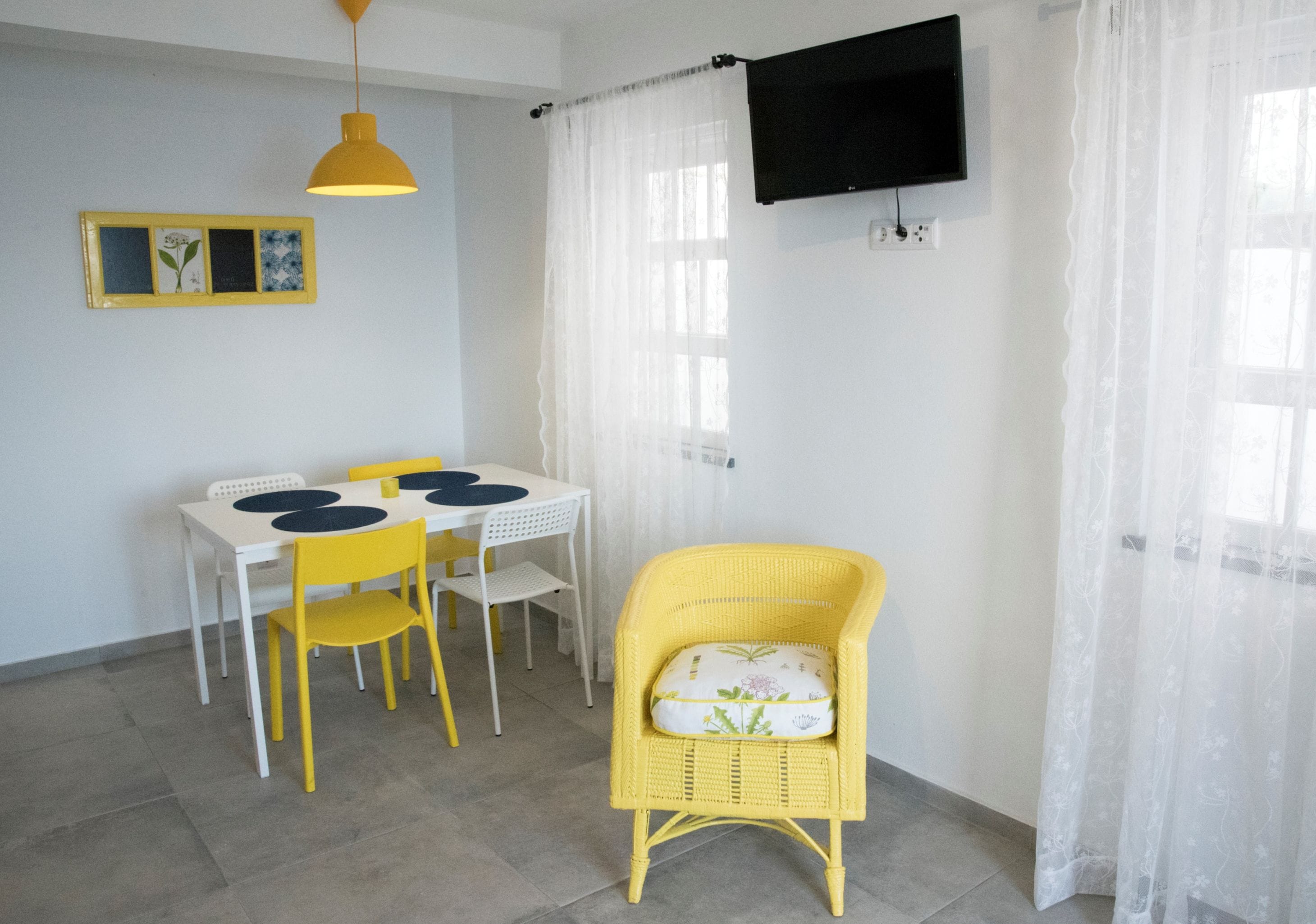 A room with a dining table, chairs, a television mounted on the wall, and a window with sheer curtains.