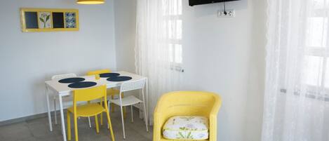 A room with a dining table, chairs, a television mounted on the wall, and a window with sheer curtains.