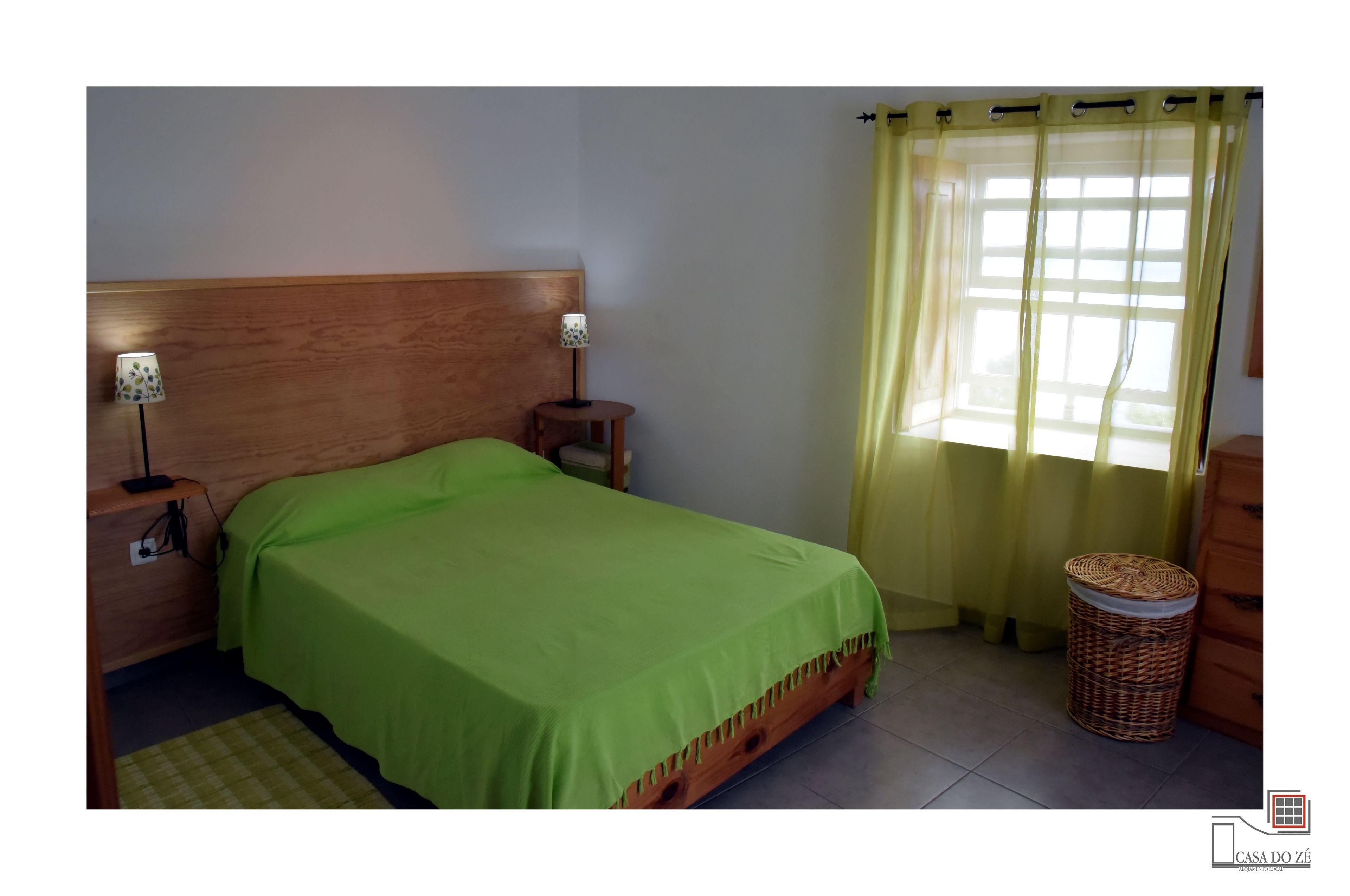 Una cama individual con cabecera de madera, una mesita de noche con lámpara, una cesta de mimbre y una ventana con cortinas amarillas.