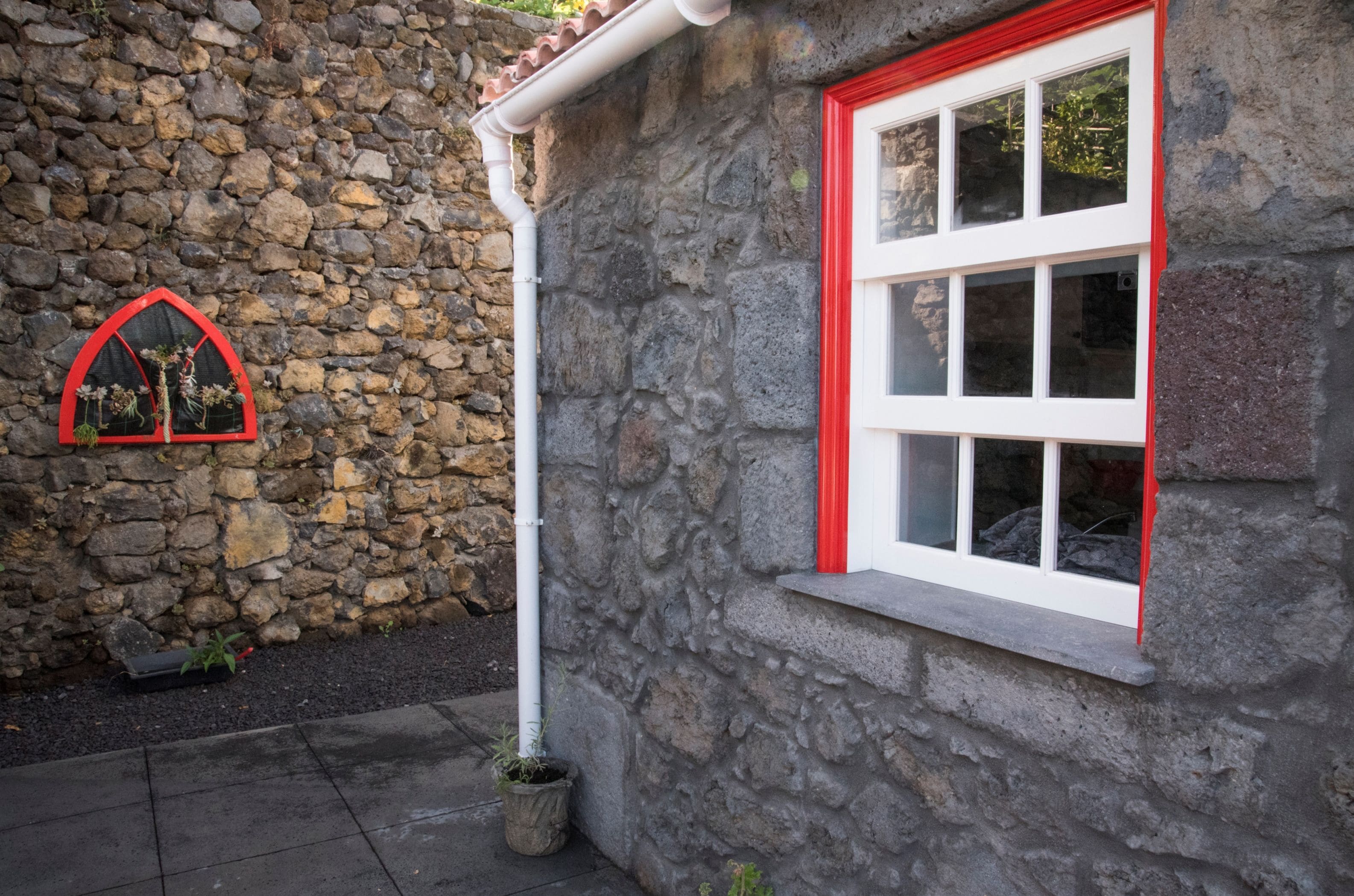 A stone building with a red-framed window and a small arched window with plants.