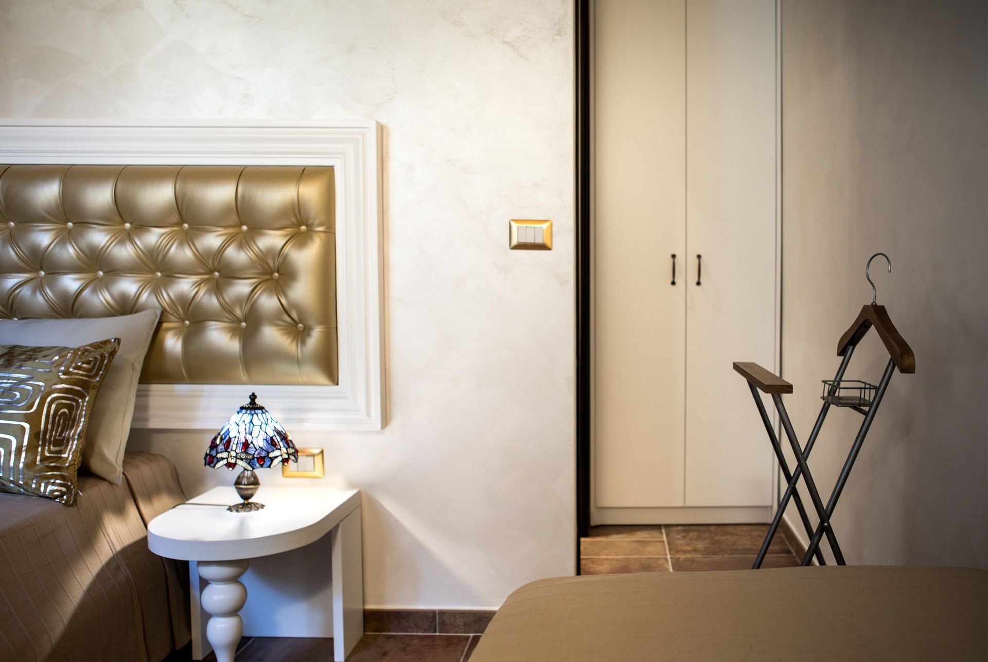 A bedroom with a tufted headboard, a bedside table with a lamp, a hanging coat rack, and a wardrobe.