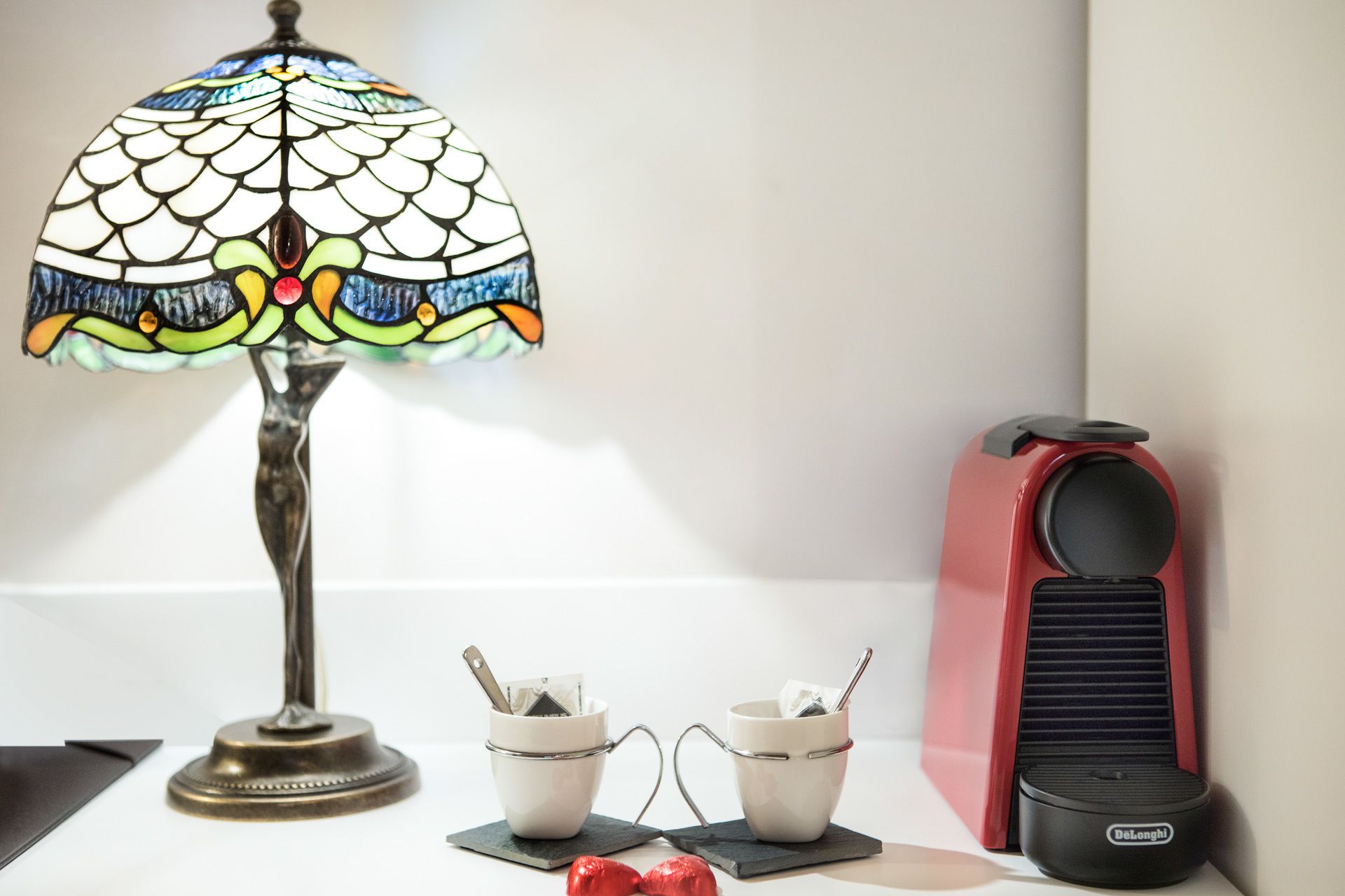 Una lámpara con pantalla de vitral, un electrodoméstico DeLonghi rojo y dos tazas con cubiertos sobre posavasos.