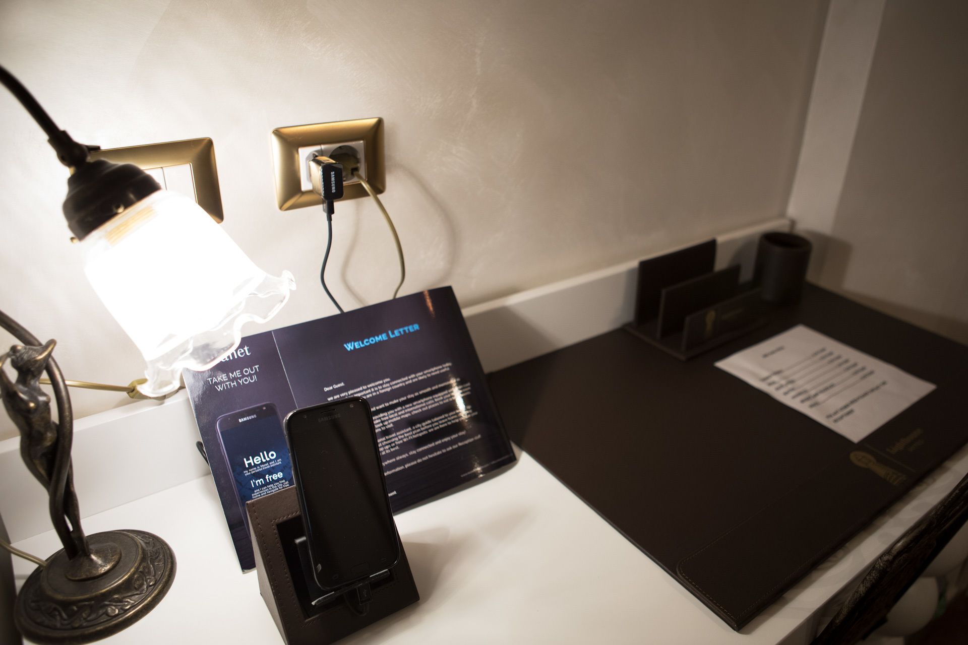 A reception desk with a Samsung phone, a welcome letter, and a lamp.