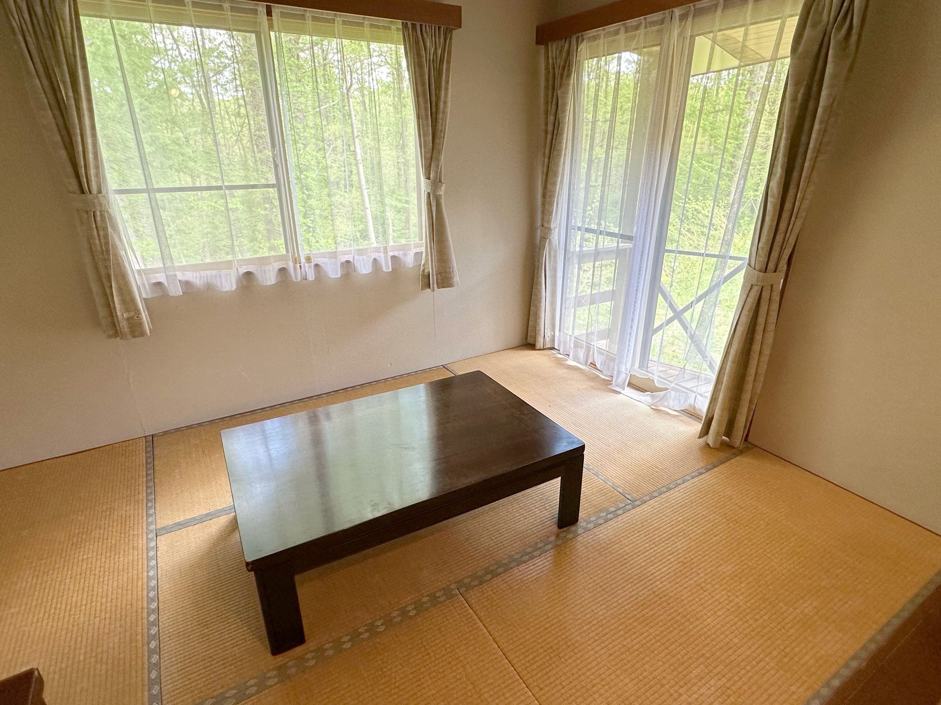 A room with tatami flooring, a wooden table, and two windows with sheer curtains.