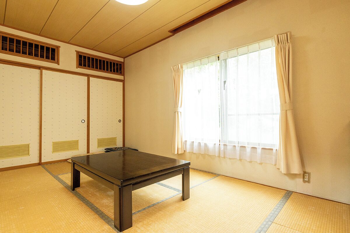 Ein traditioneller japanischer Raum mit Tatami-Bodenbelag, einem niedrigen Tisch, Schiebetüren und einem Fenster mit Vorhängen.