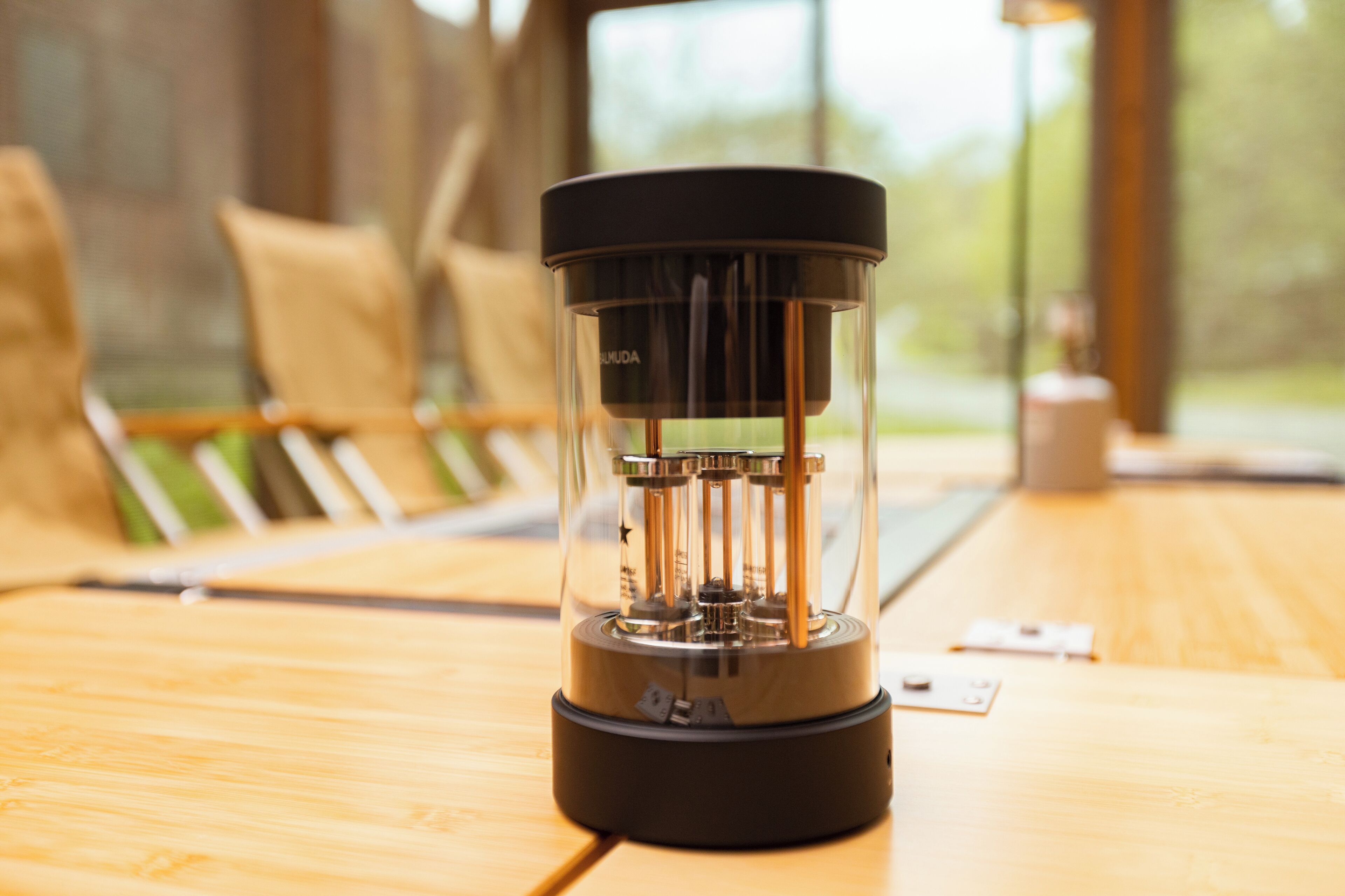 A transparent coffee maker on a wooden table with a window view in the background.
