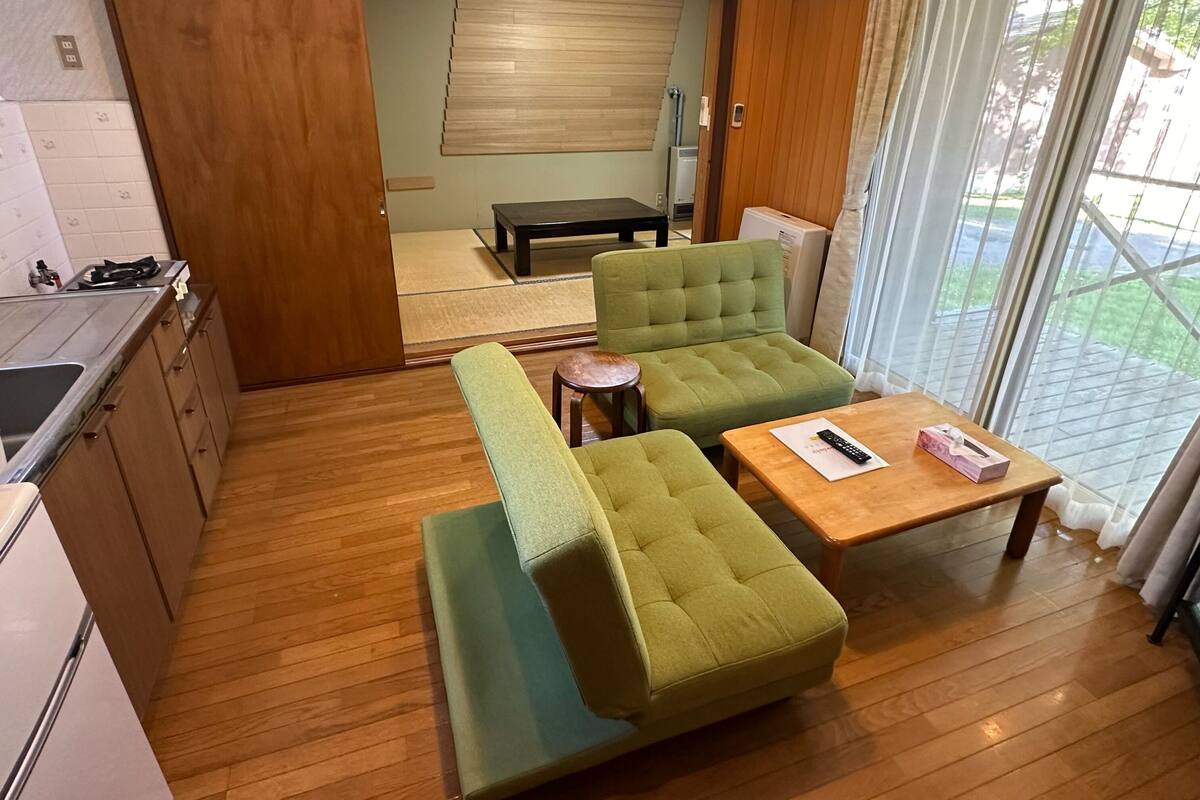 A compact living space with a kitchenette, a green sofa, a small table, and a view through a sliding door.